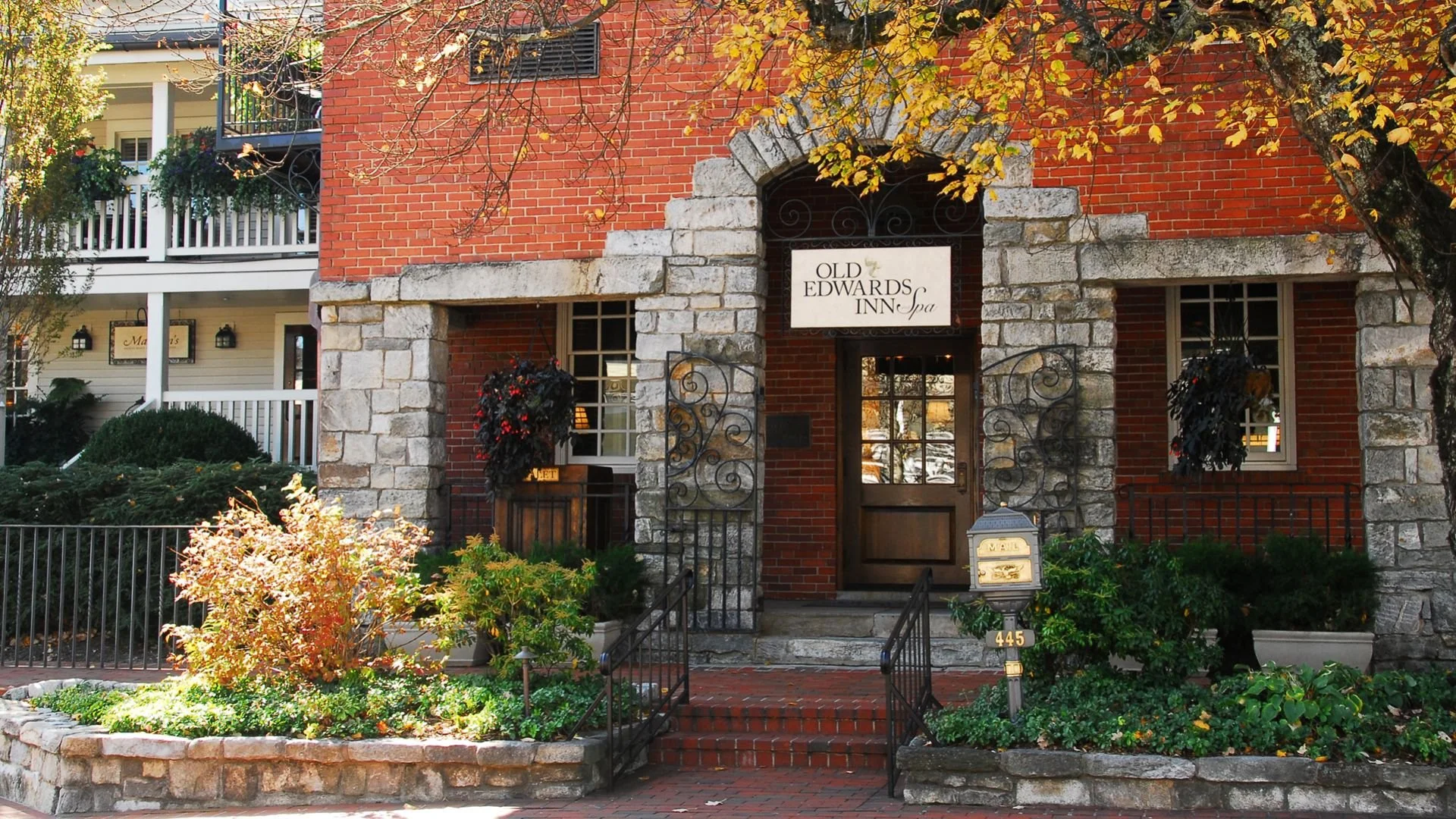 The exterior of the Old Edwards Inn & Spa, a multi-story brick building with stone accents, featuring a front garden with plants and a small brick staircase leading to the entrance, surrounded by trees with autumn leaves.