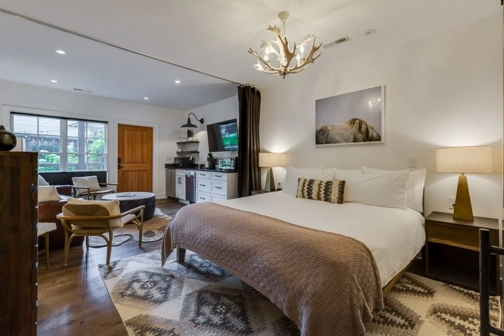 Modern hotel room with a large bed, nightstands, lamps, artwork, a seating area with chairs and a table, and a television.