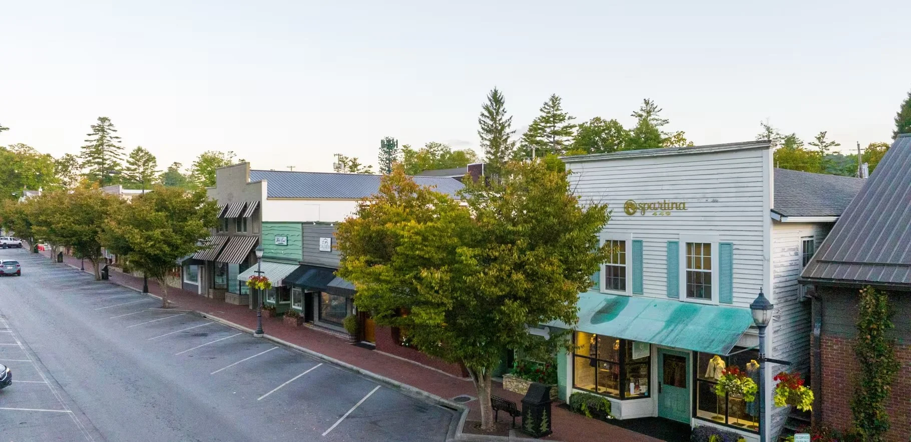 A small-town street with storefronts, trees with autumn leaves, parked cars, and a lamp post.