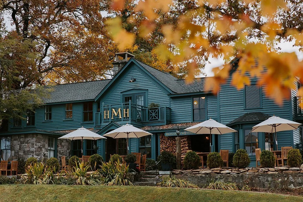 A large blue house with multiple windows, a stone foundation, and a small balcony with the initials 'H M H' on it. There are three white umbrellas and outdoor seating in front of the house, surrounded by trees with orange and yellow fall leaves.