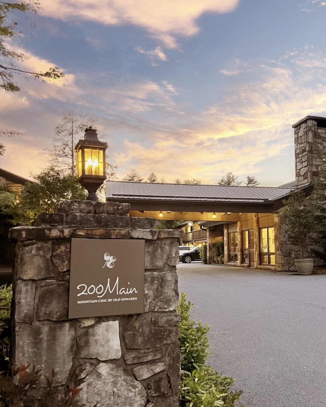 A stone sign with a butterfly logo and the text '200 Main Mountain Chic by Old Edwards' in front of a lodge-style building with yellow lighting, surrounded by trees during sunset.