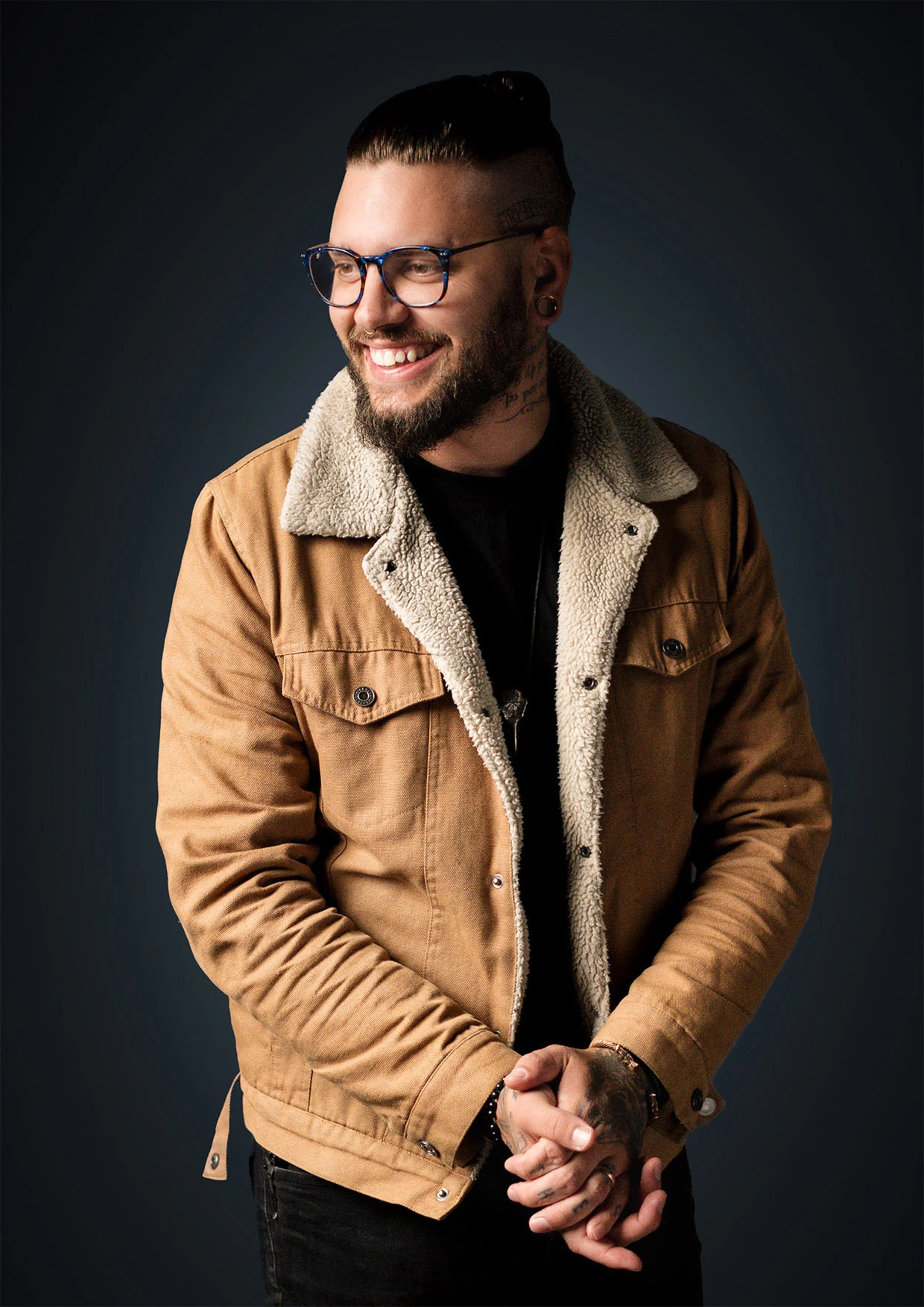 Man smiling, wearing glasses, a brown jacket with a fleece-lined collar, and black pants, against a dark background.