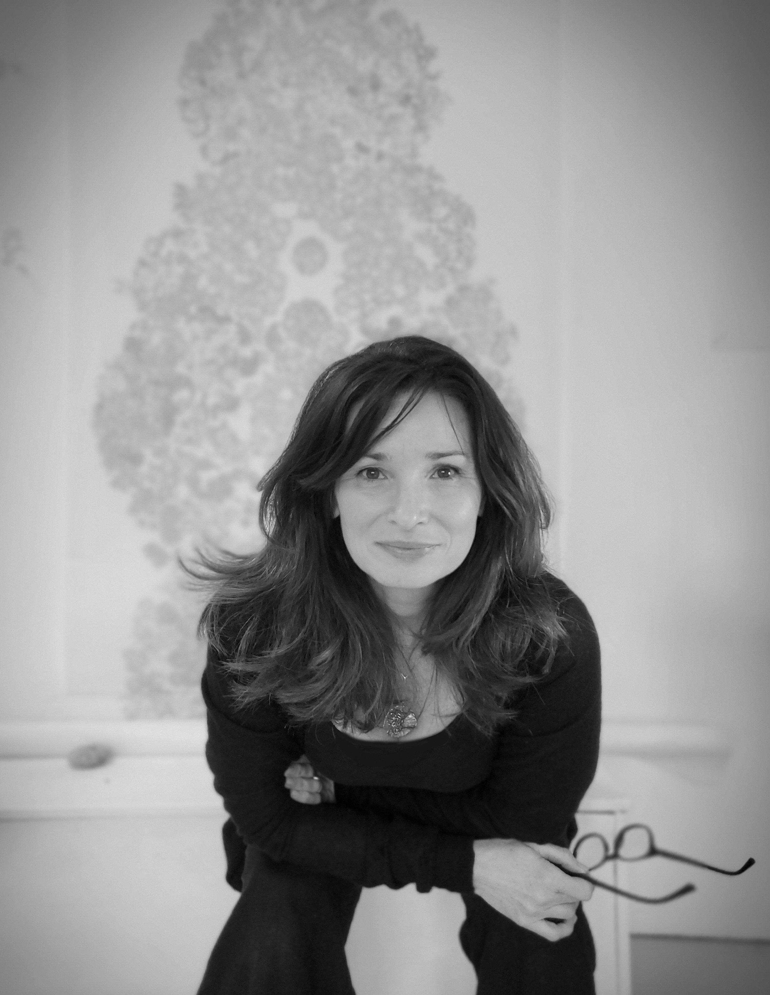 Black and white photo of the artist with long wavy hair, smiling and holding glasses, sitting or crouching in front of a blurred background with plant based motifs.