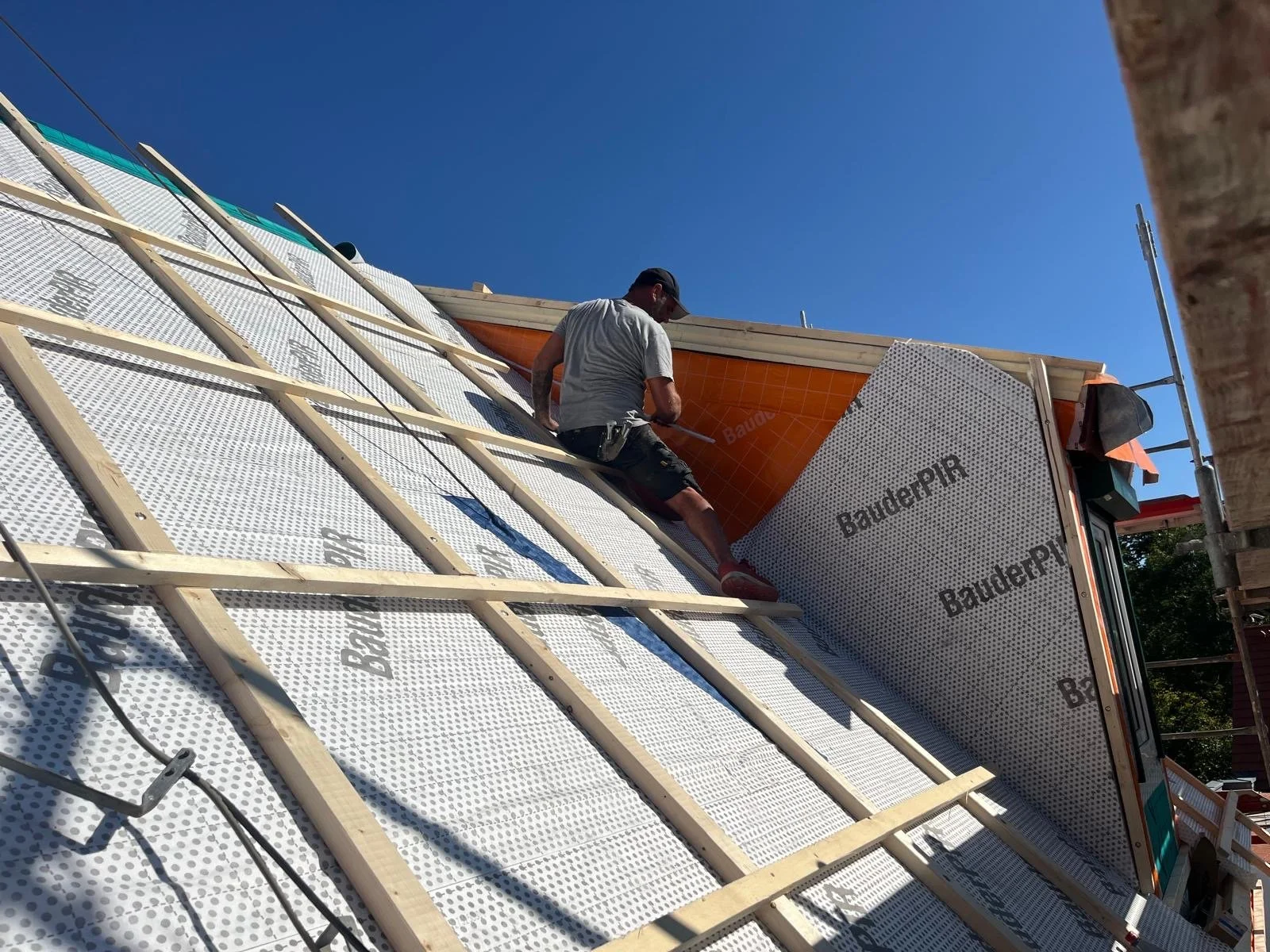 Ein Handwerker installiert eine Dacheindeckung auf einem Hausdach bei strahlendem Sonnenschein.