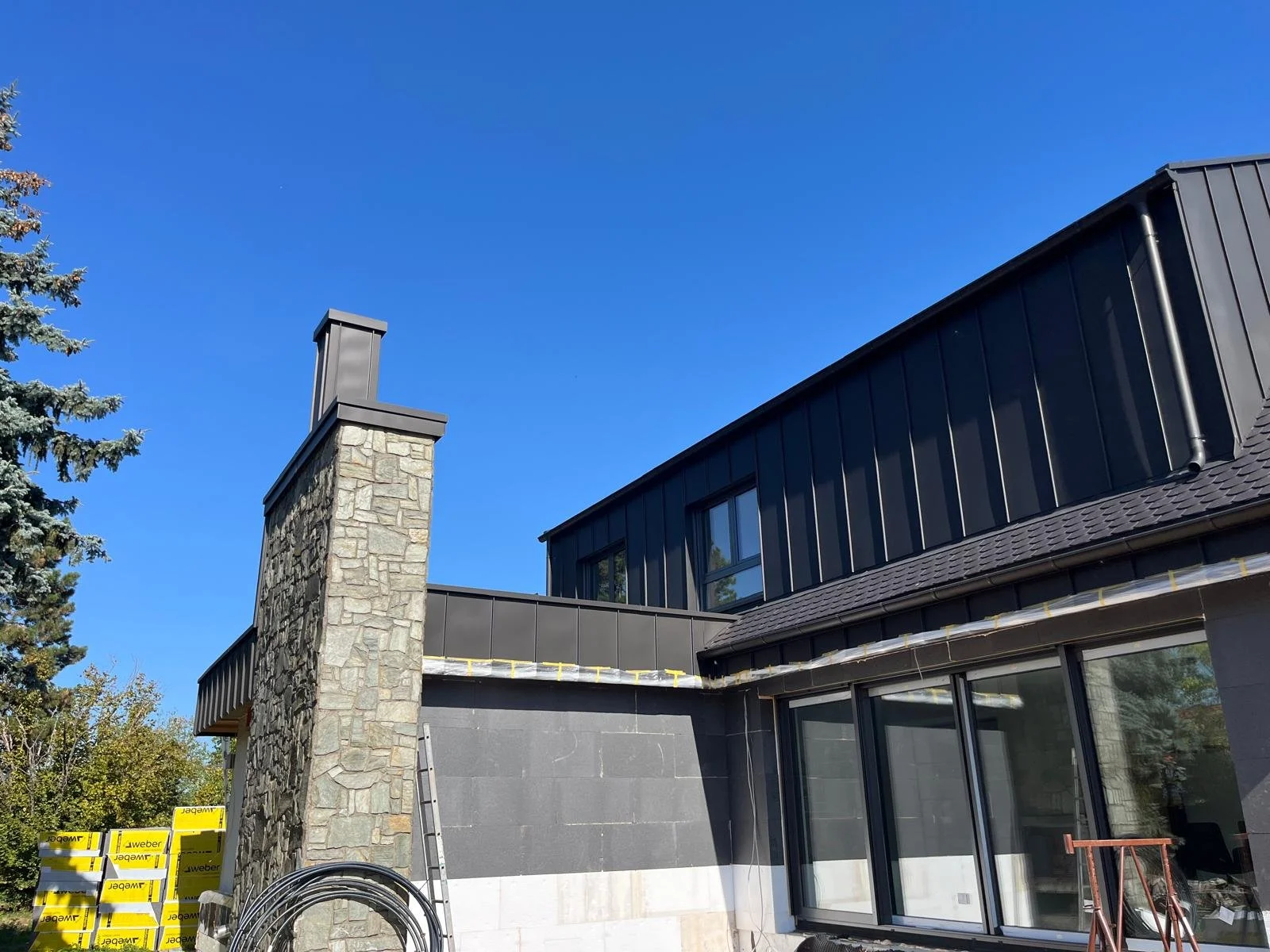 Außenansicht eines modernen Hauses mit dunkler Fassade, großen Fenstern, und einem Schornstein aus Naturstein unter einem klaren blauen Himmel.