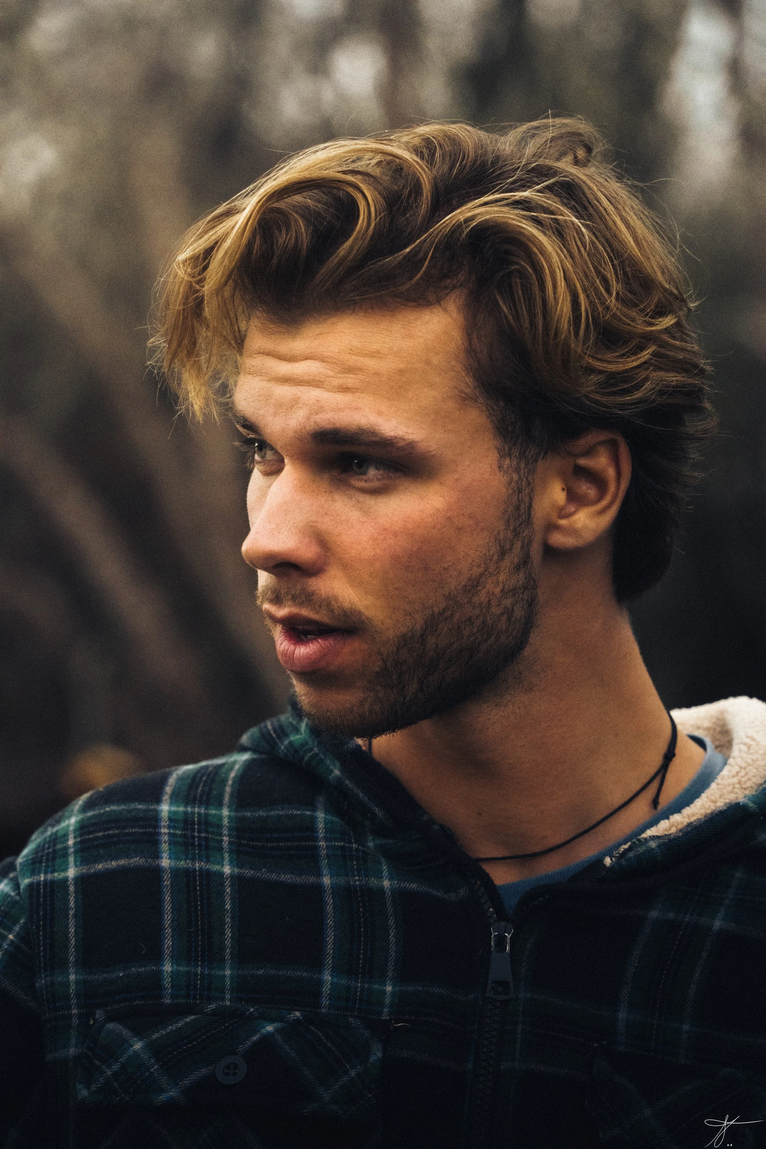 Close-up of a young man with wavy, light brown hair, a mustache, and beard, wearing a plaid jacket, in an outdoor setting with blurred background.