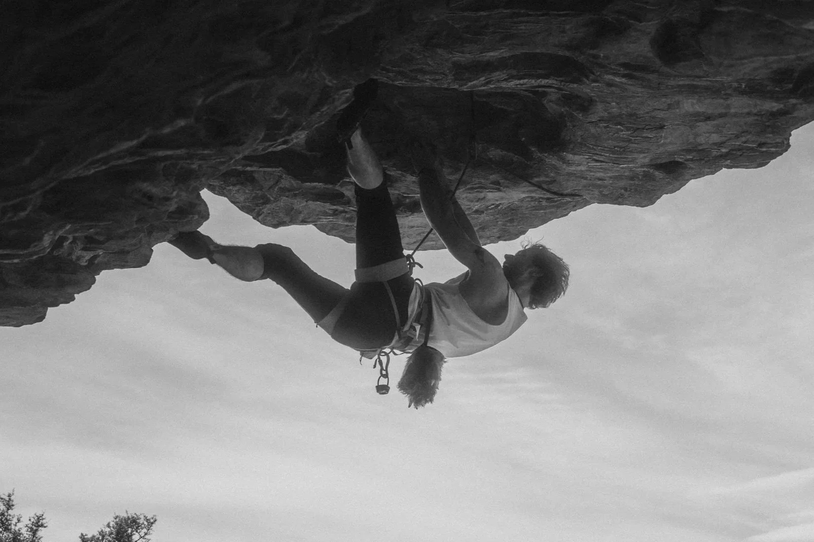 Person rock climbing on a steep outdoor rock face, wearing a tank top, shorts, and climbing shoes, with a clouded sky in the background.
