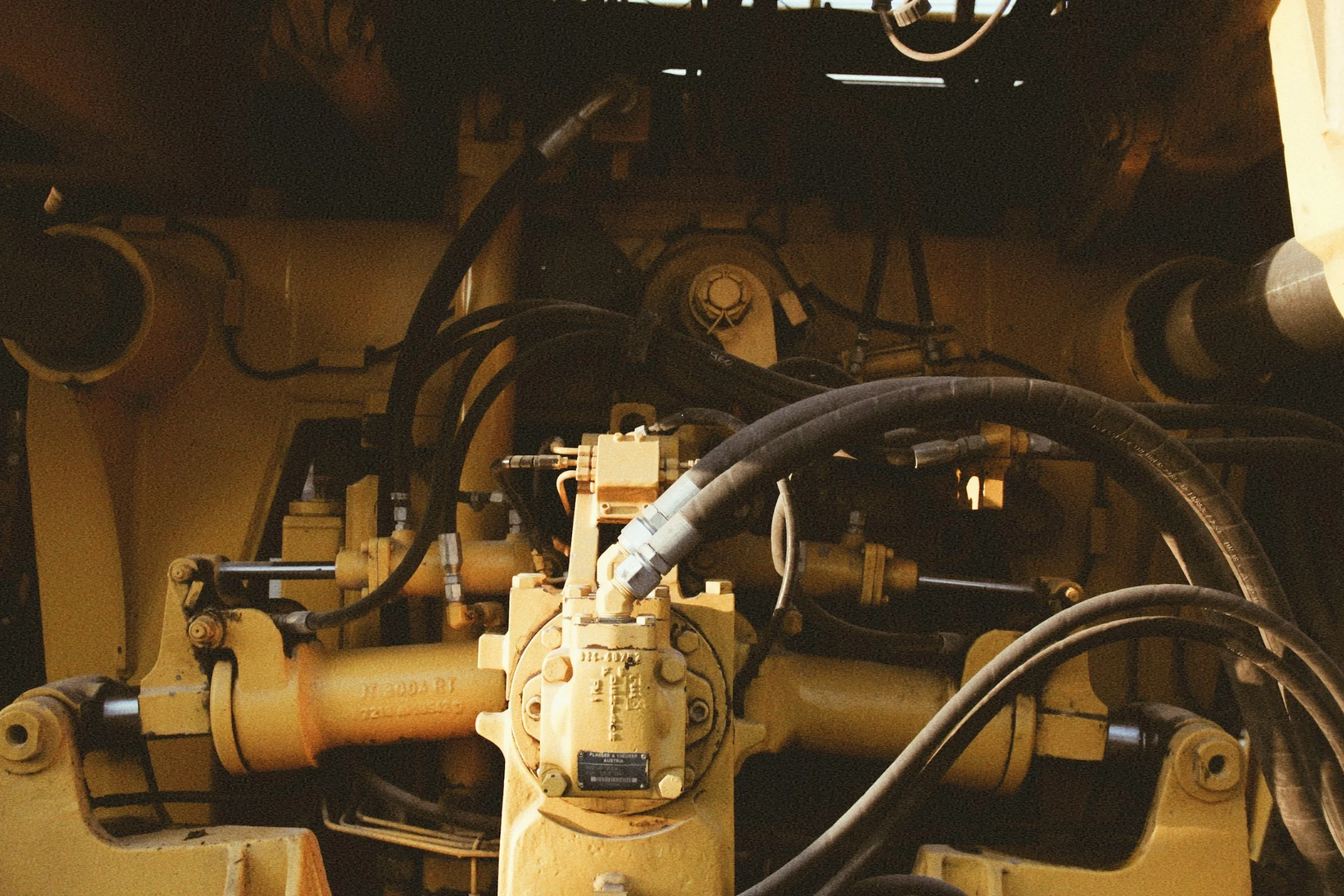 Close-up of the machinery's hydraulic system with yellow painted metal components, hydraulic hoses, and fittings inside a construction or industrial vehicle.