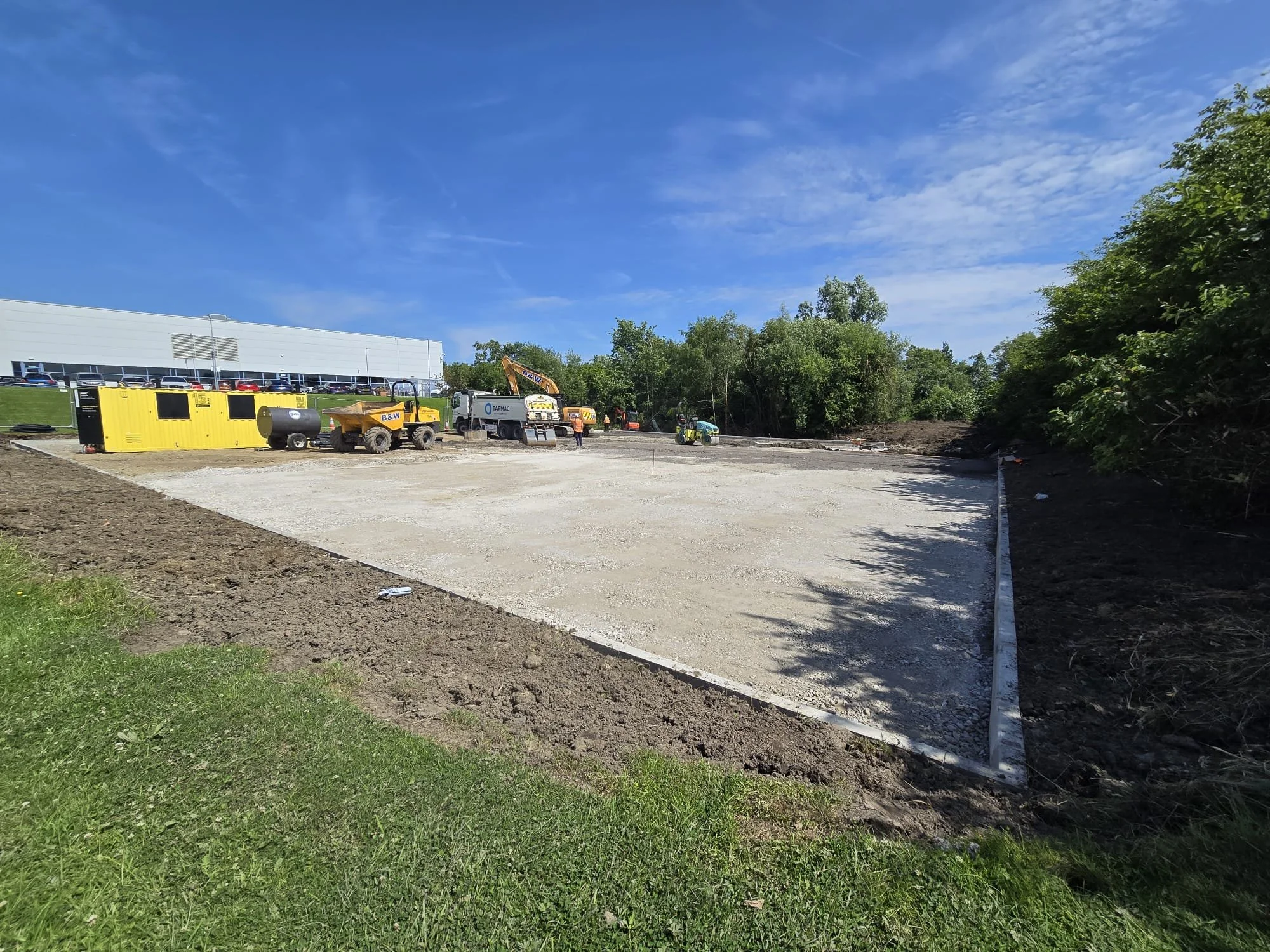 Construction site with a gravel lot, construction equipment, and trees in the background under a blue sky.