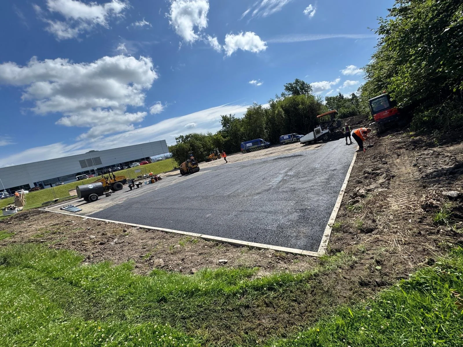 Workers laying down asphalt on a parking lot with construction equipment and vehicles nearby, under a partly cloudy sky.