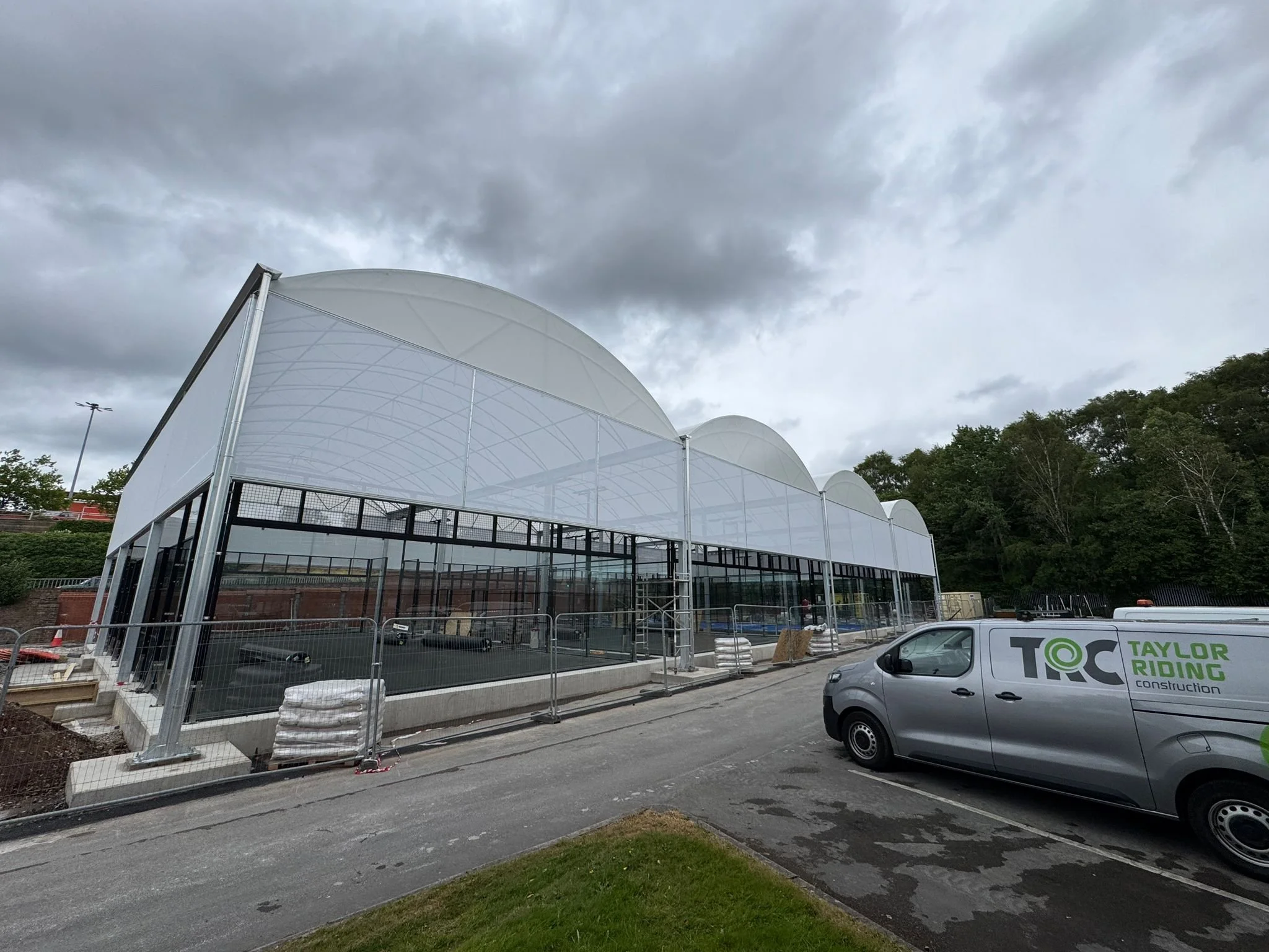 A large construction site with a greenhouse-like structure under construction, surrounded by fencing, with a gray Taylor Riding construction van parked nearby. The sky is overcast with dark clouds, and there are trees in the background.