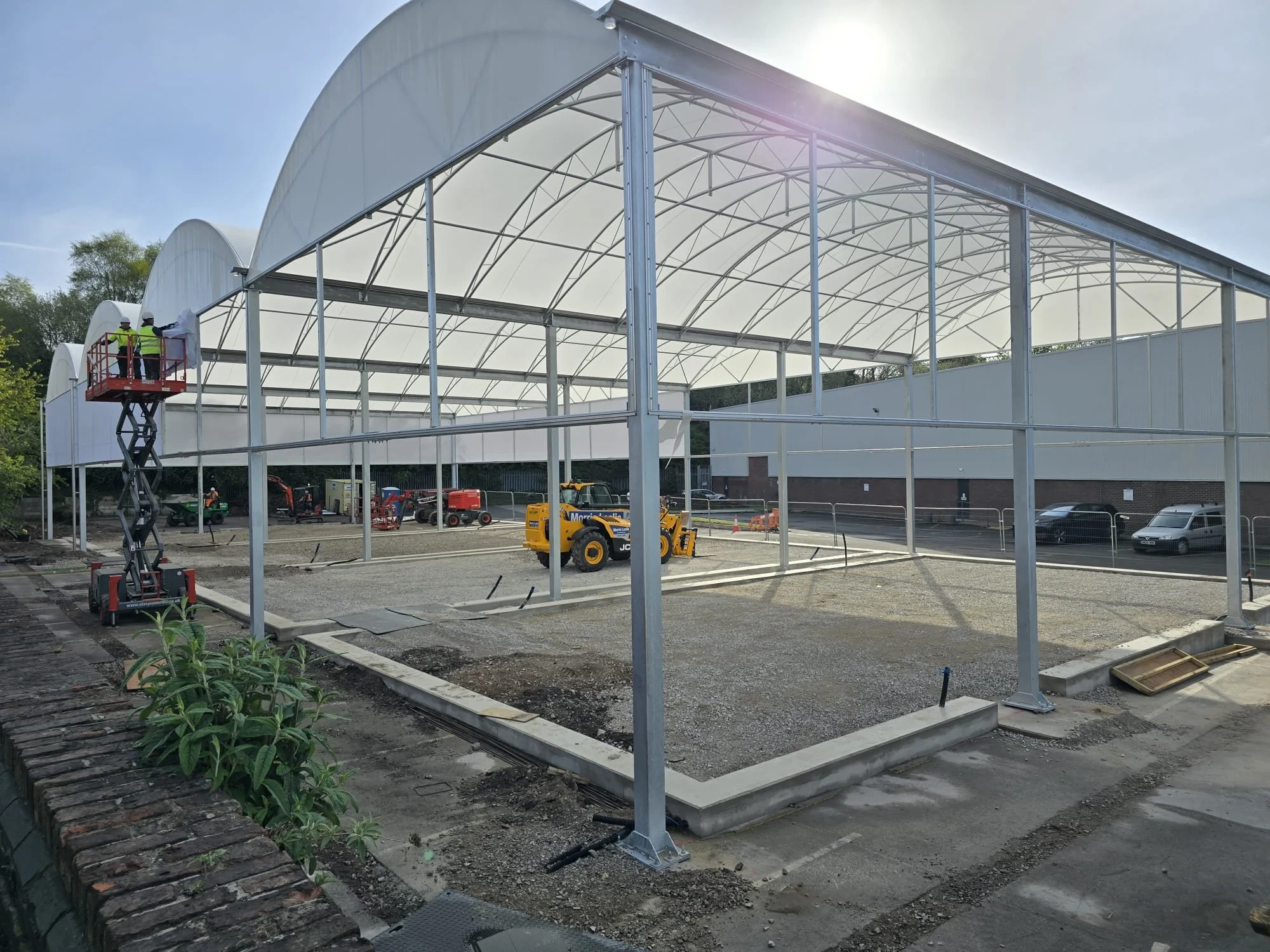 Construction of a large greenhouse with a metal framework and a white arched roof, with workers on a lift and construction vehicles on site.