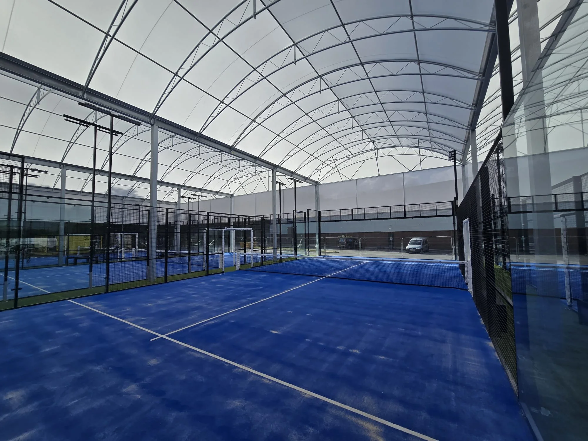 Indoor tennis court with blue surface, enclosed by mesh fences, under a large curved, translucent roof.