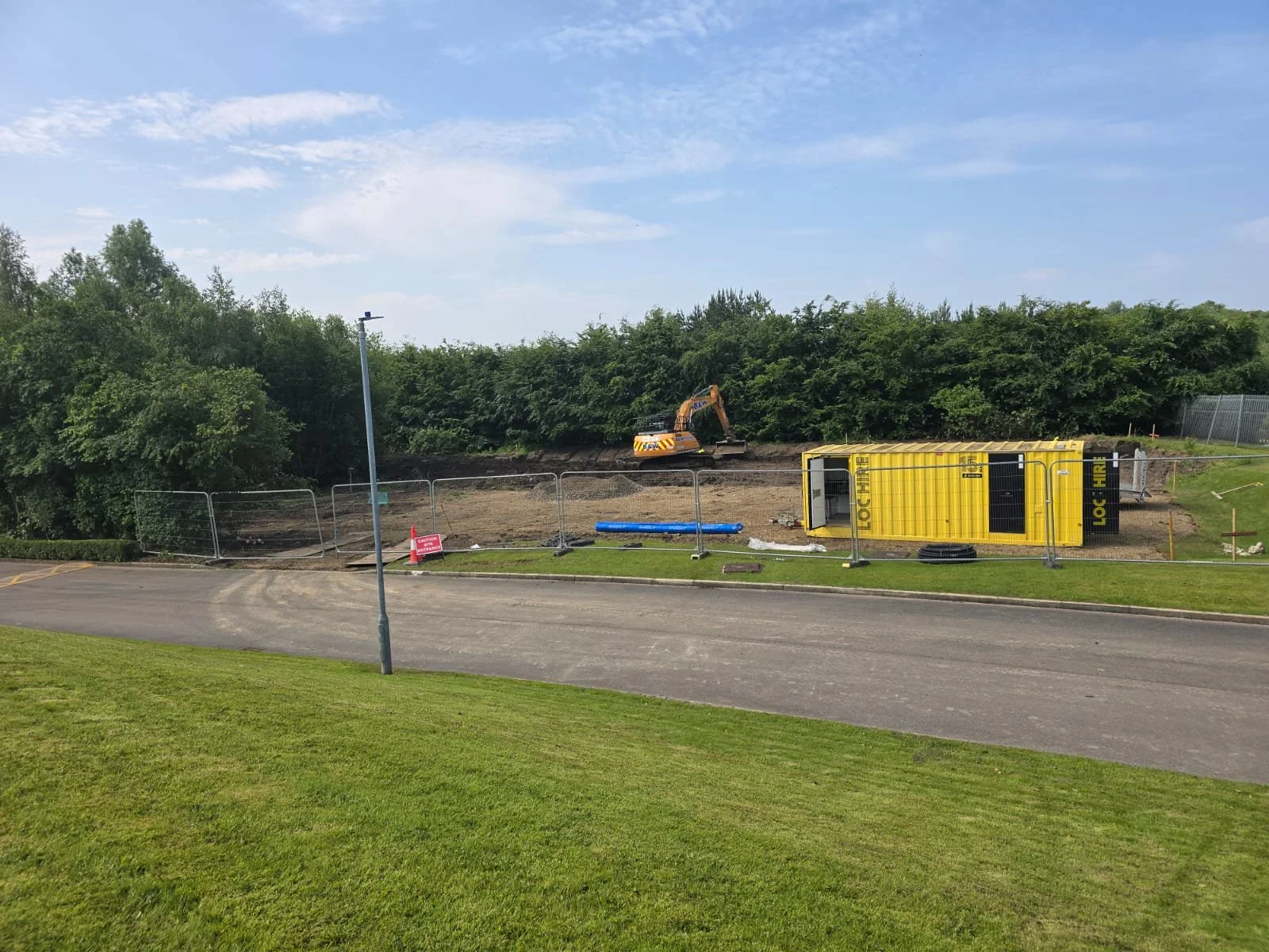 Construction site with a toppled yellow shipping container, a yellow excavator behind it, and a dirt area enclosed by a temporary fence, next to a parking lot and sidewalk with grass.