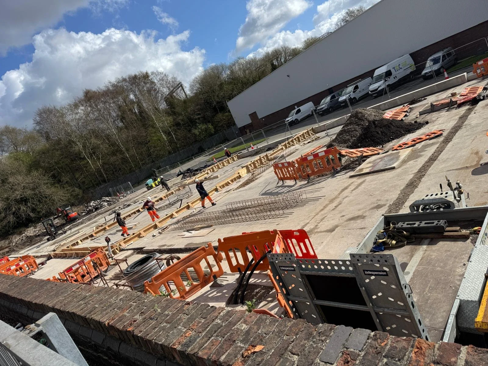 Construction workers working on a building site with wooden frames, orange safety barriers, and construction materials, alongside parked vehicles and a hill with trees under a partly cloudy sky.