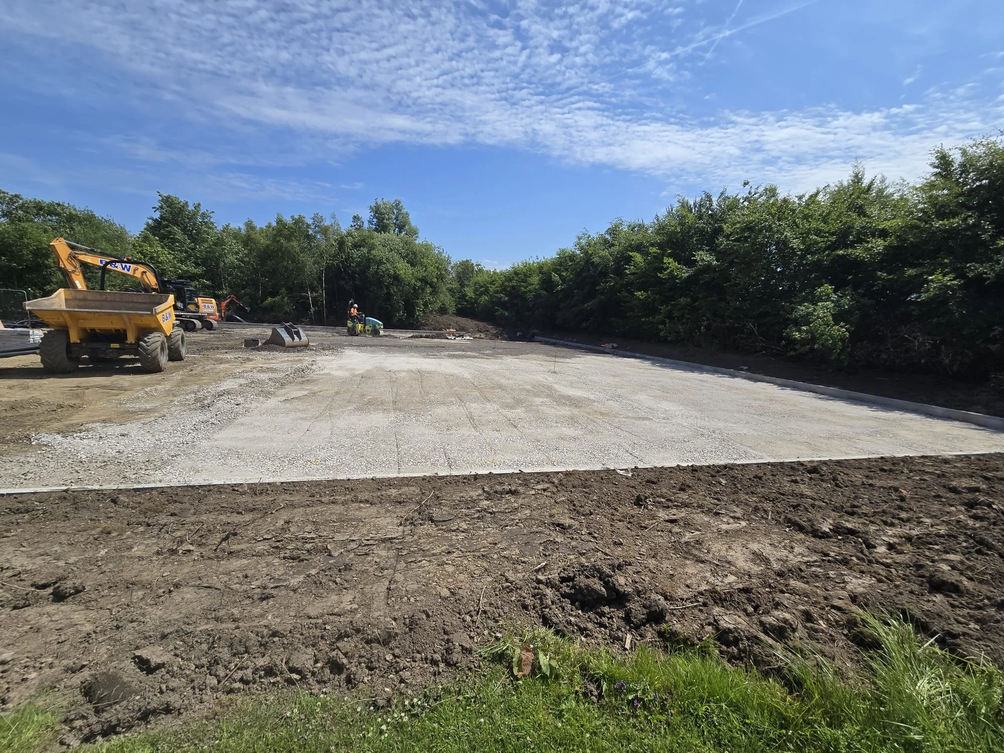 Construction site with machines and a worker, ground being prepared for paving, surrounded by trees and a partly cloudy sky.