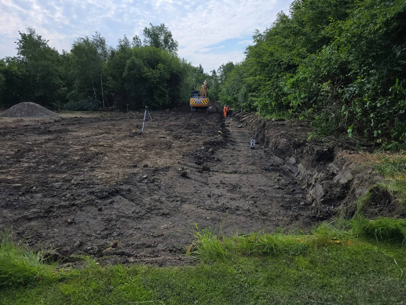 Construction site with excavator and workers in a muddy area surrounded by trees.