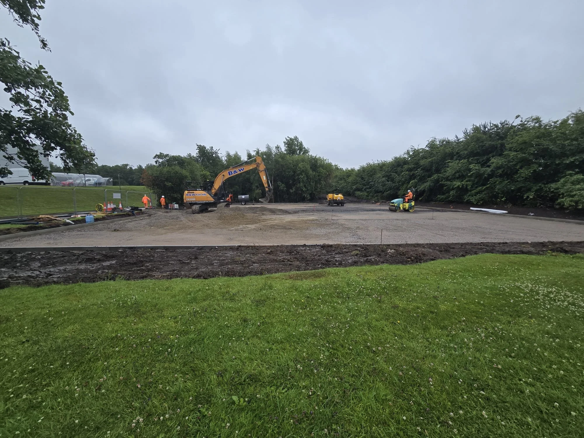 Construction site with workers, excavator, and road roller on a cloudy day, surrounded by greenery.