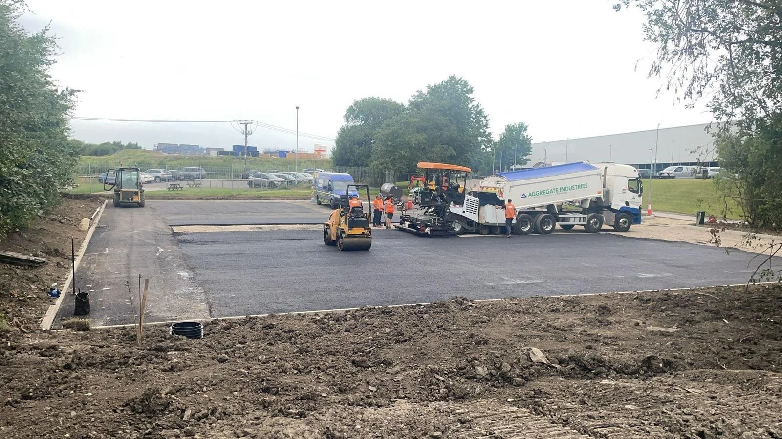 Construction workers are paving a parking lot with asphalt using heavy machinery, including a roller compactor and an asphalt paver.