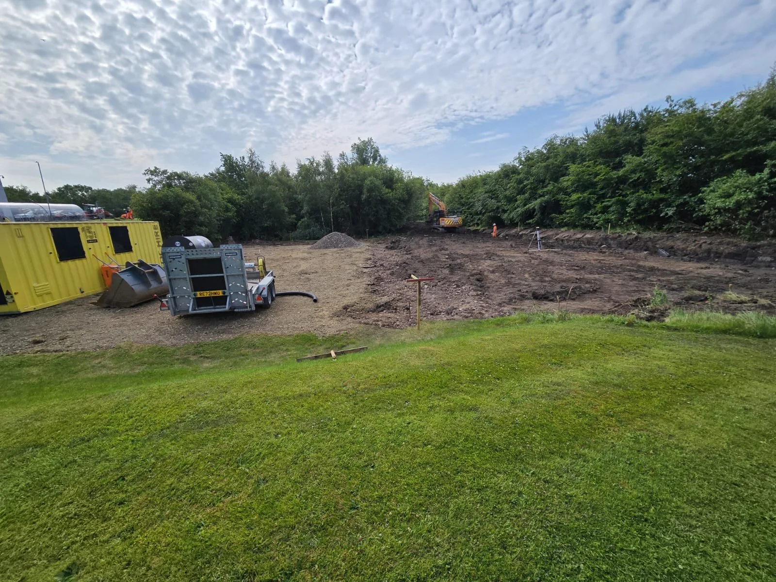 Construction site with machinery, yellow portable toilets, and a cleared area of dirt, surrounded by trees under a partly cloudy sky.