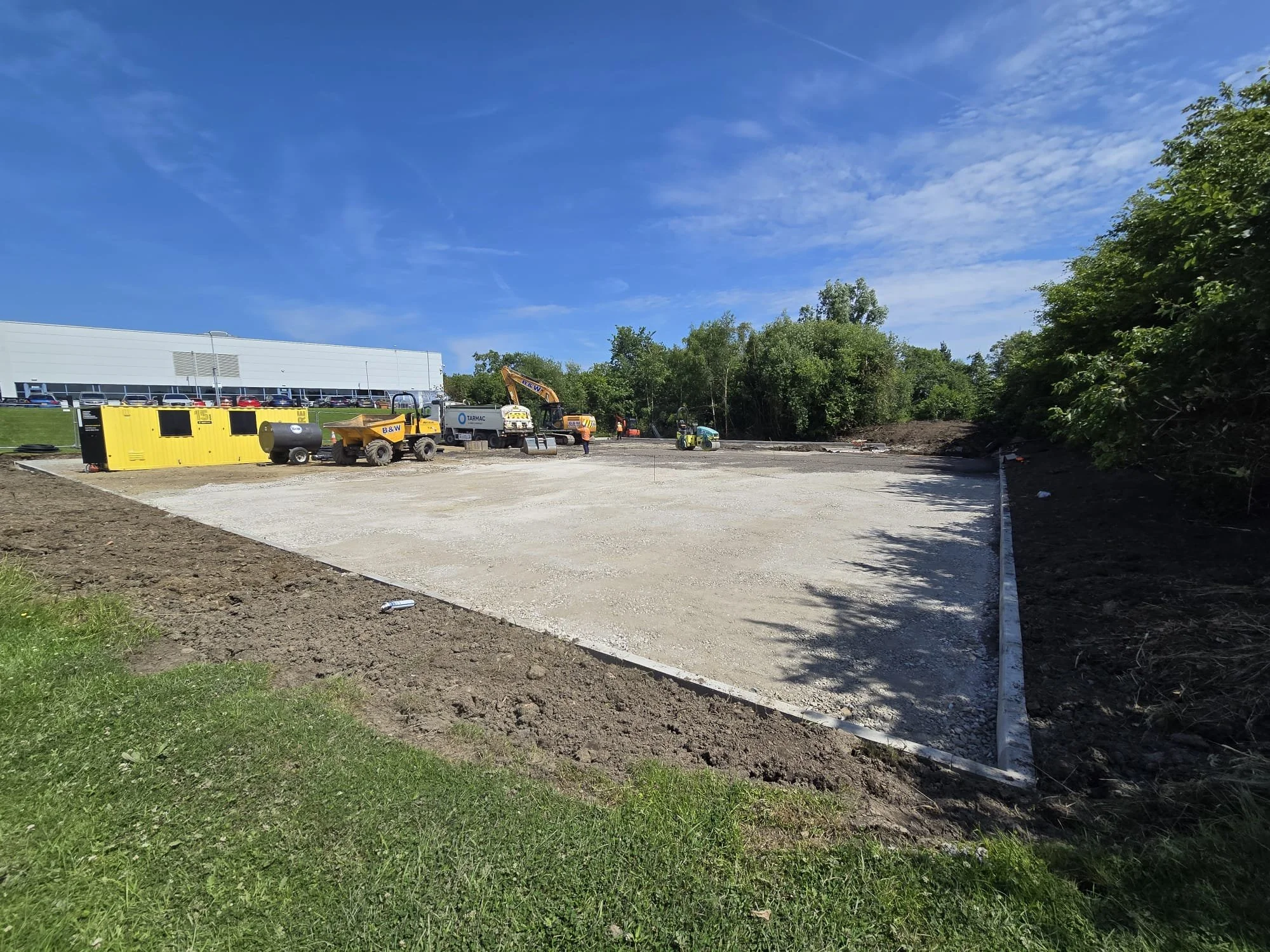 Construction site with equipment, trucks, and workers on a cleared area, bordered by grass and trees under a blue sky.