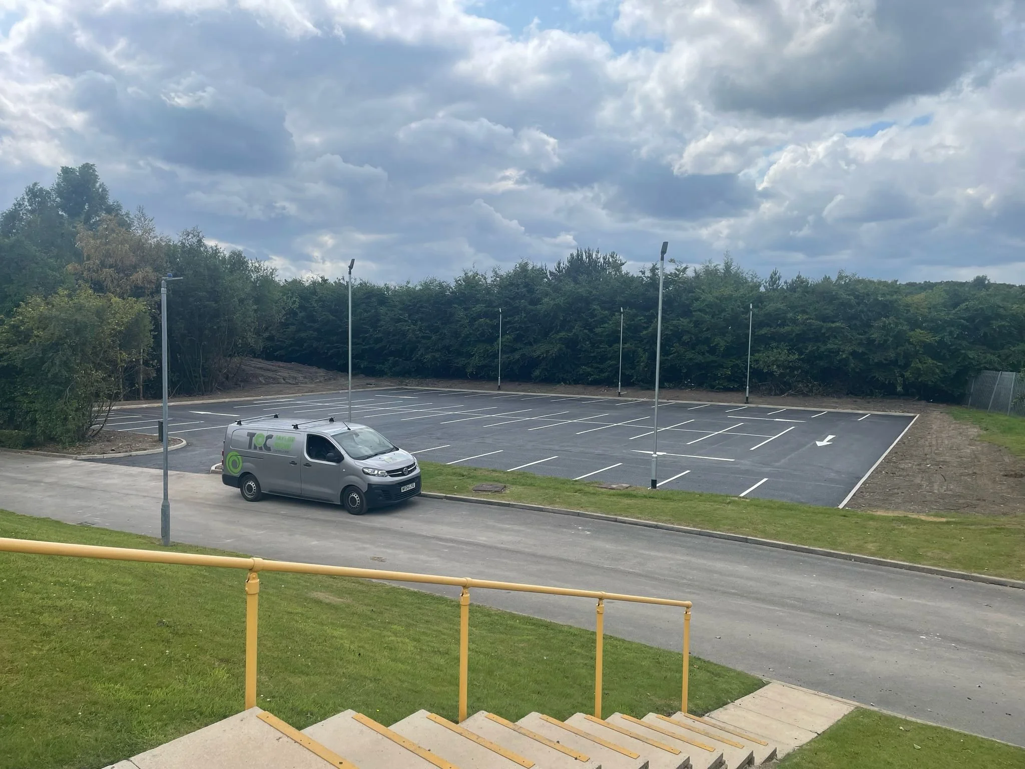 Empty parking lot with a few streetlights and a silver van parked near yellow handrails, surrounded by trees and bushes under a partly cloudy sky.