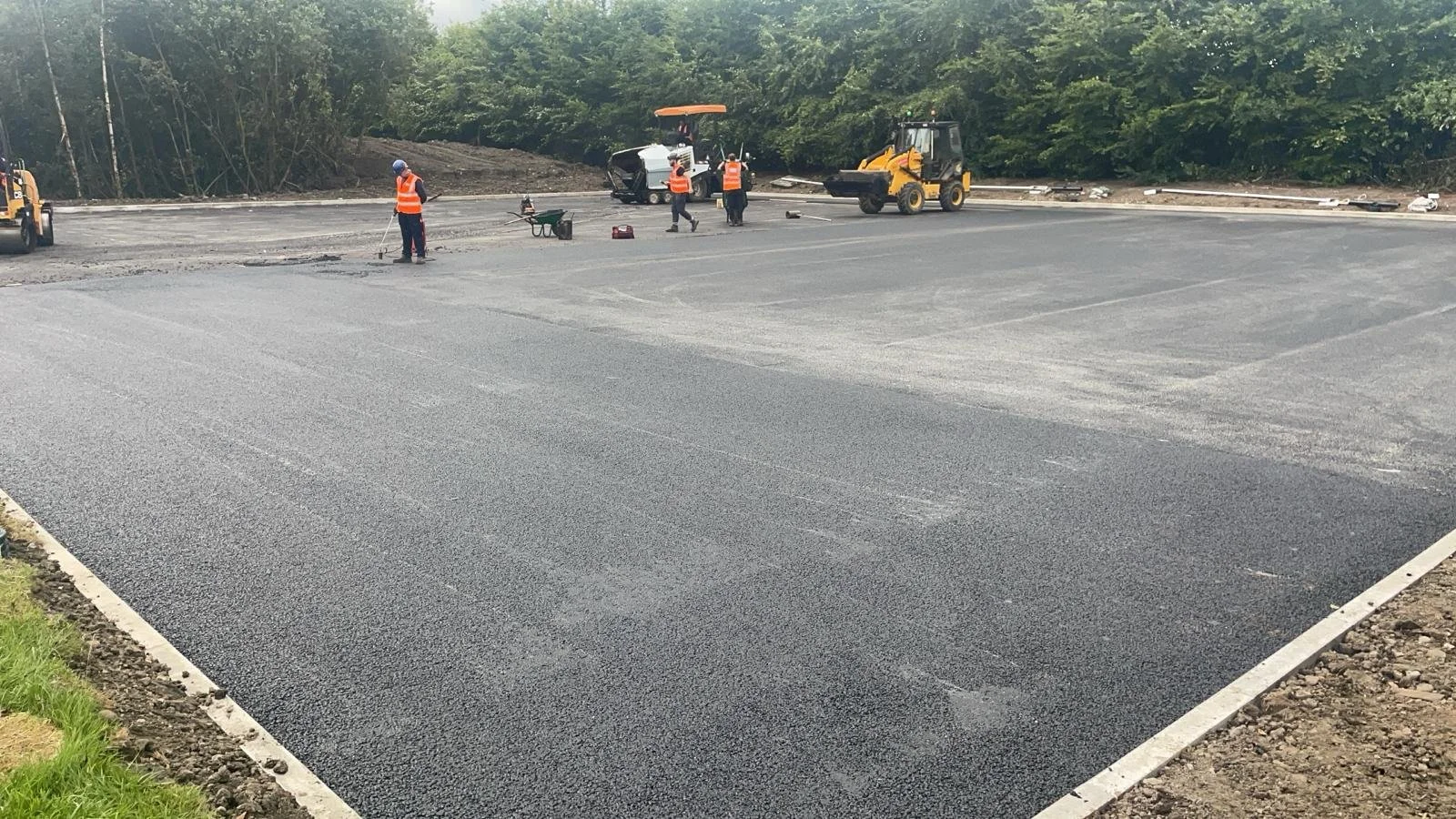 Workers in orange safety vests and helmets working on paving and resurfacing a parking lot, with construction equipment and vehicles in the background.