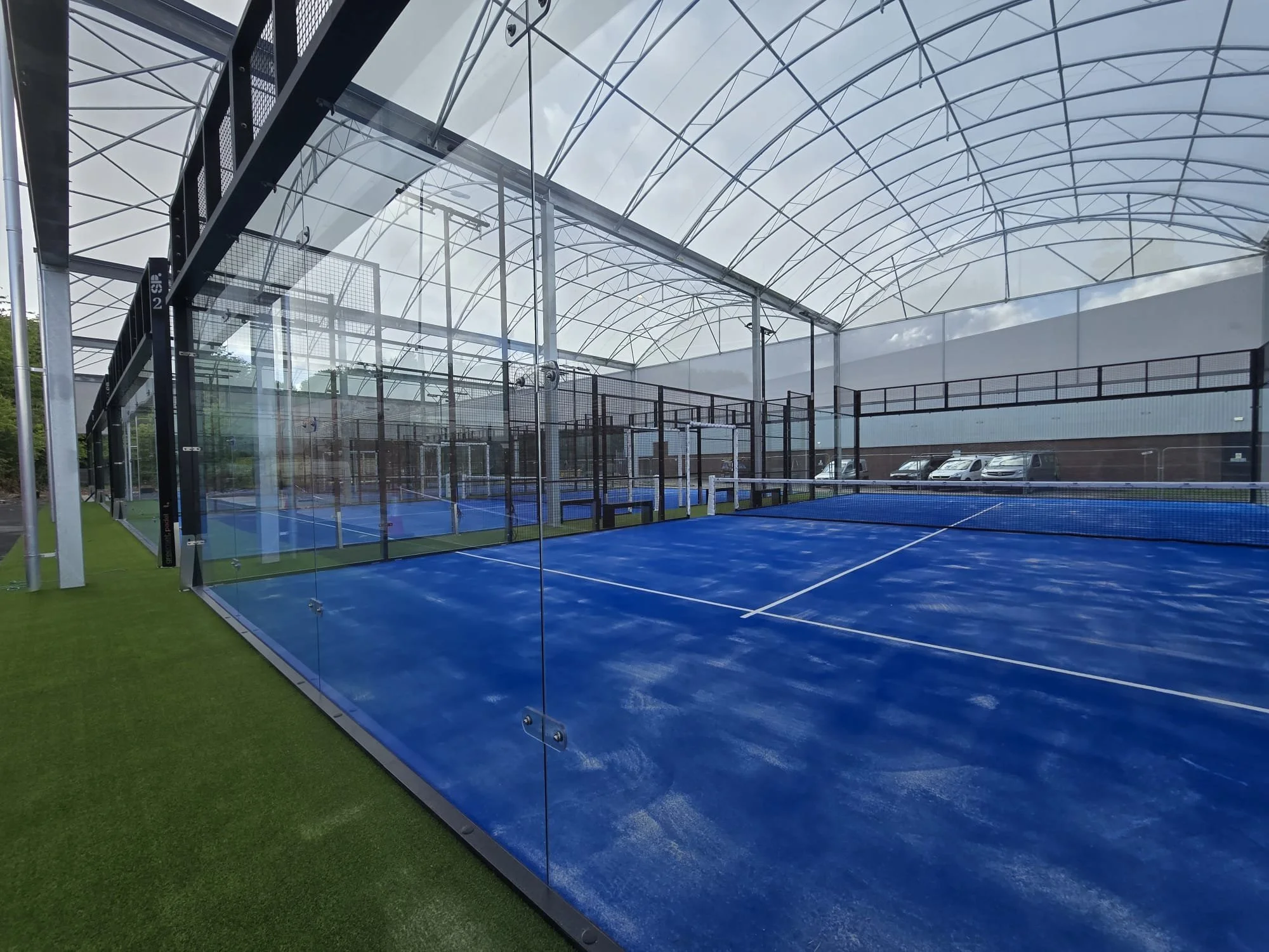 Indoor tennis court with blue surface, enclosed by glass walls, in a large climate-controlled structure with a white arched roof.