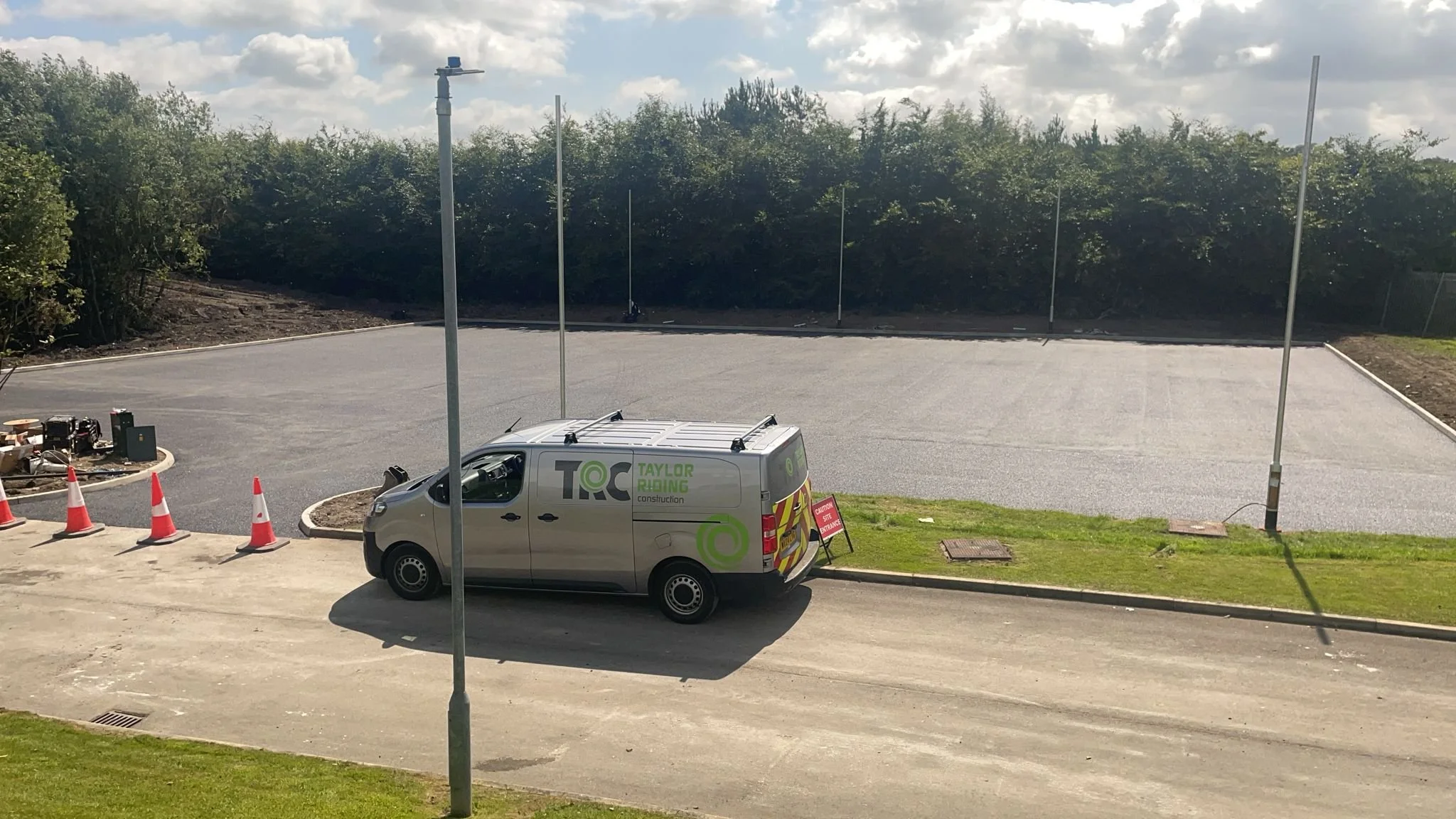 Construction site with a gray van parked on a dirt and asphalt road, orange traffic cones, and a large empty paved area in the background with trees and a cloudy sky.
