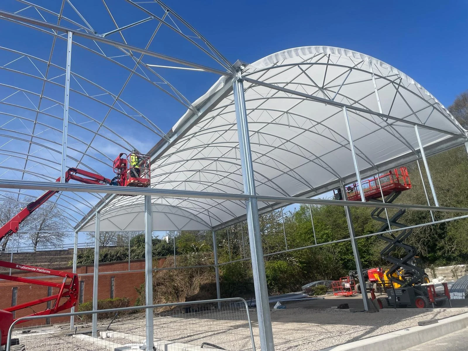 Construction workers on lifts installing a large, white, arched greenhouse structure with metal framework under a clear blue sky.