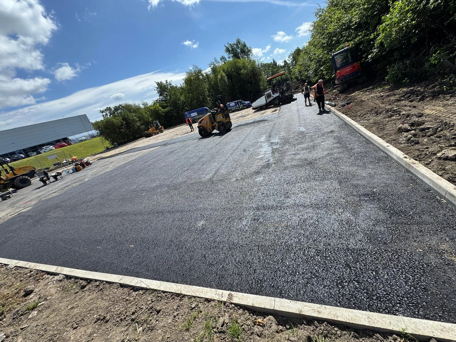 Construction workers pave asphalt on a parking lot, with construction equipment and trees in the background, under a partly cloudy sky.