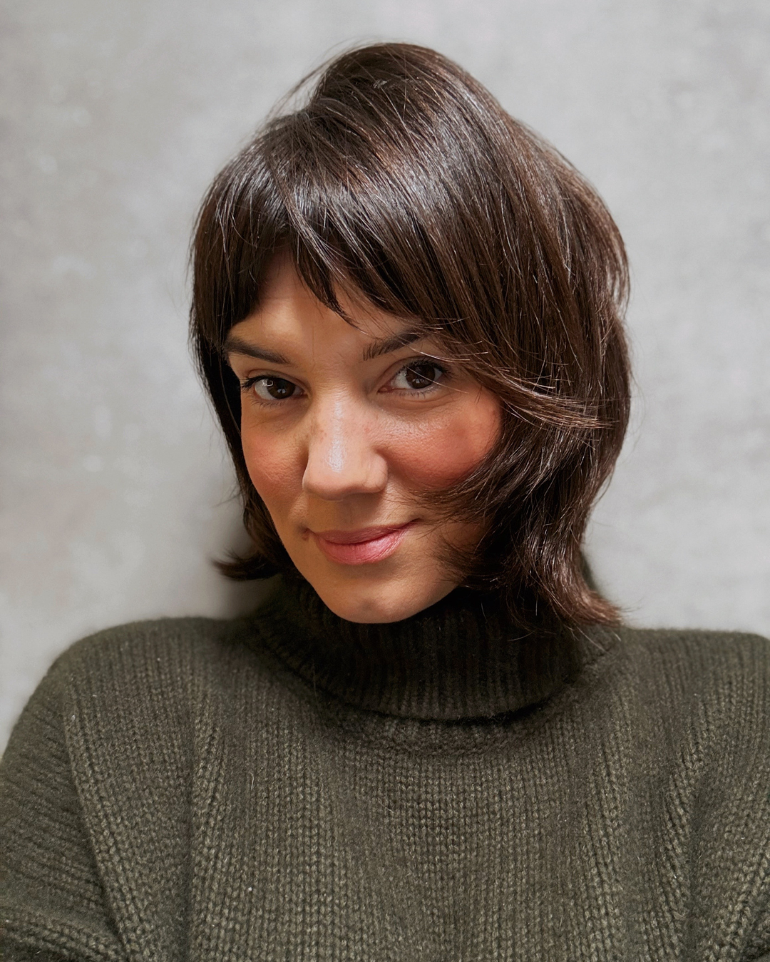 Jeune femme avec coupe de cheveux brune coiffée en dégradé, portant un pull vert foncé, souriant légèrement devant un mur gris.