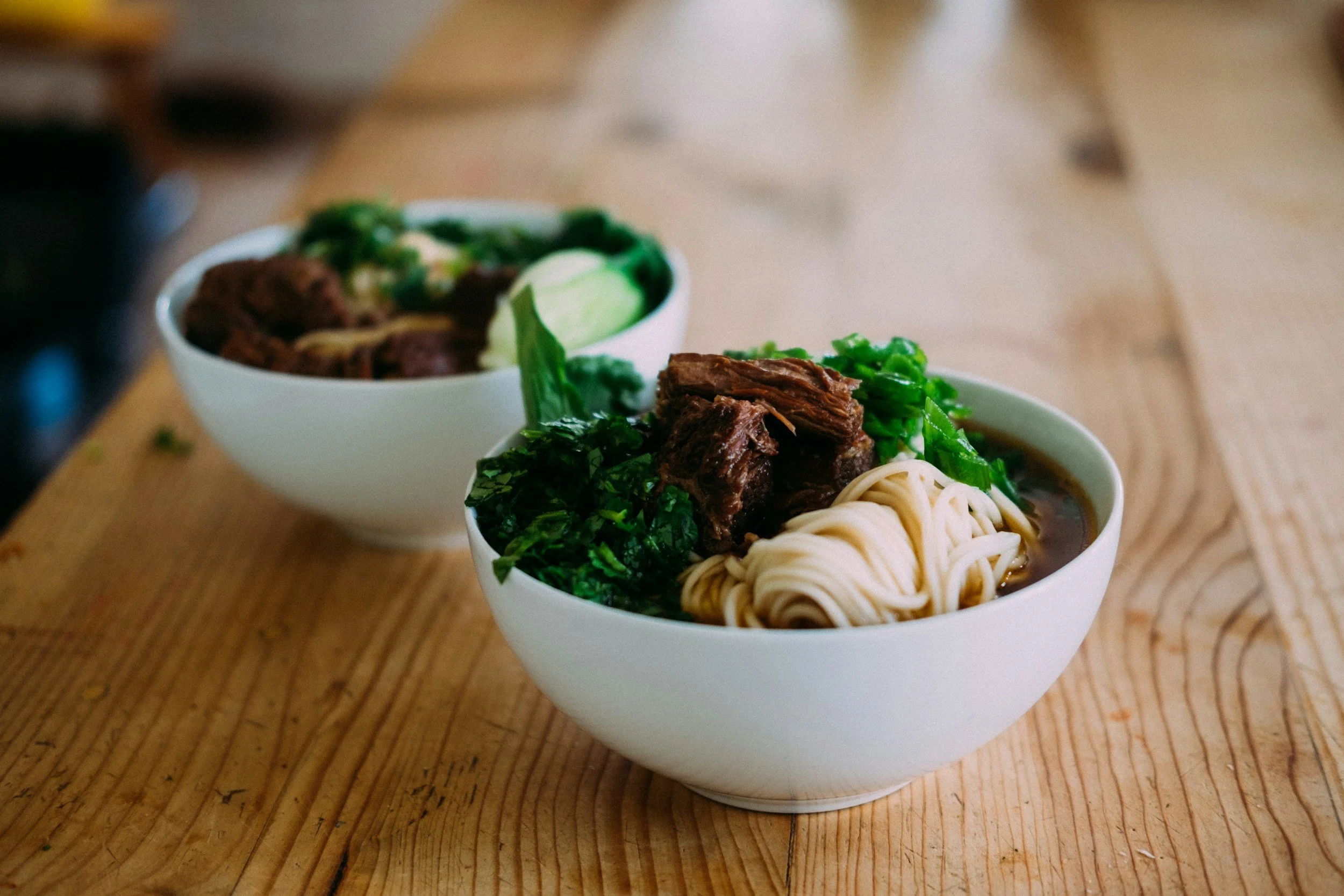 Bol de ramen avec viande, légumes verts et nouilles, en premier plan, et un autre bol à l'arrière sur une table en bois.
