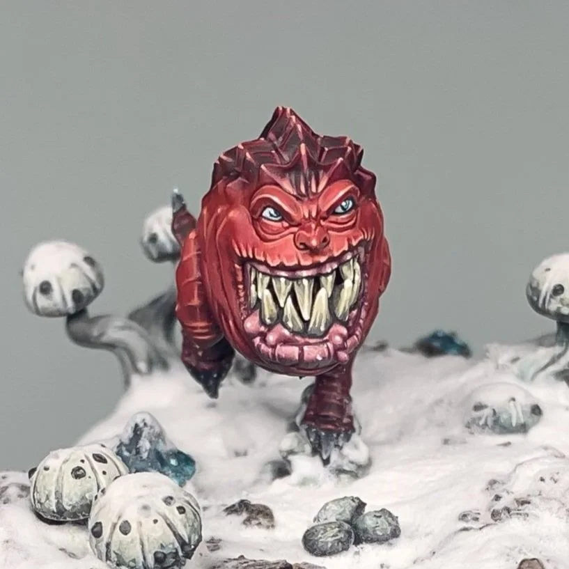 Une figurine de créature monstrueuse rouge avec un visage anthropomorphe, squig aux dents pointues, sur socle enneigé.