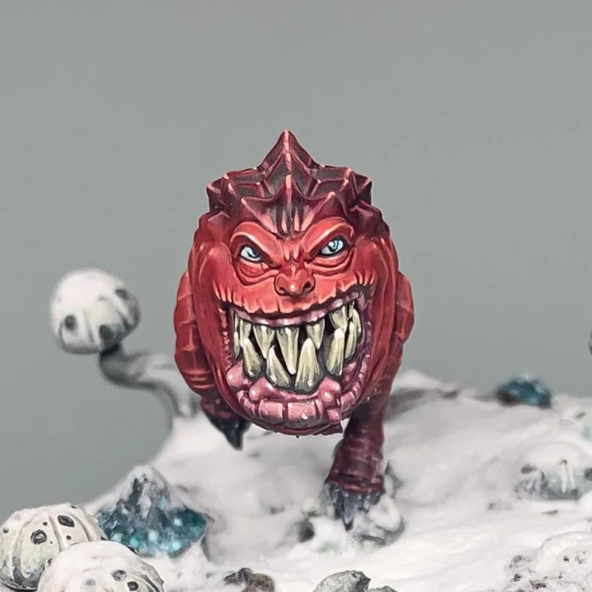 Une figurine de créature monstrueuse avec un visage rouge, des yeux perçants et une bouche pleine de dents pointues, sur un fond blanc et socle enneigé. Squig souriant