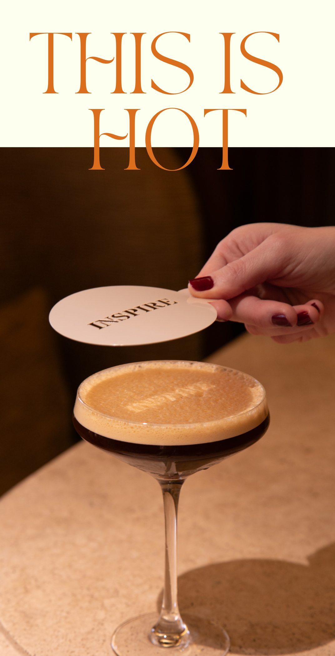 Un verre à cocktail avec une boisson foncée, une main tenant une carte avec le mot 'INSPIRE', et en haut le texte 'THIS IS HOT' en lettres orange.