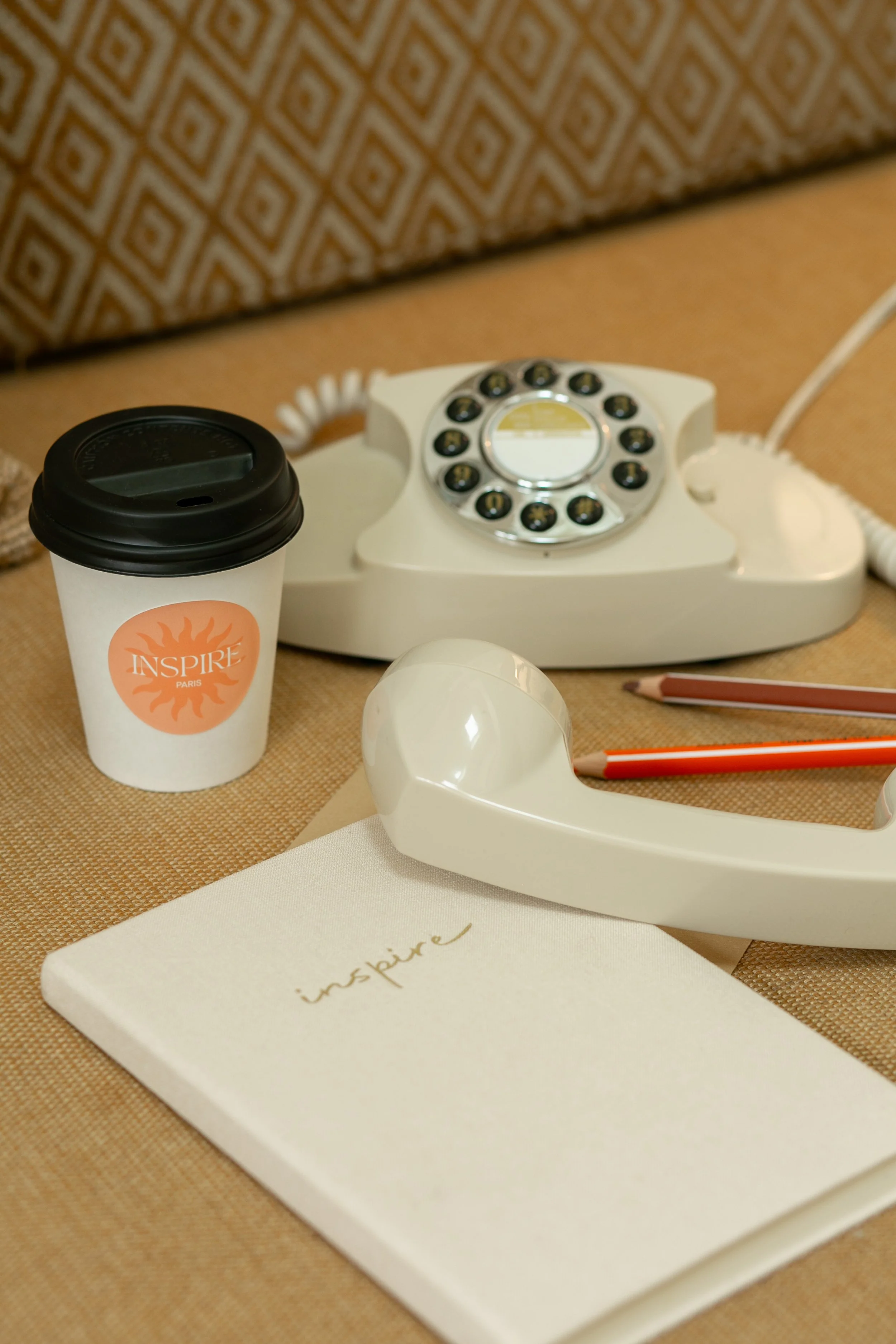 Une tasse de café, un téléphone ancien à cadran, un carnet avec l'inscription 'inspire' et deux crayons sur une surface en tissu beige.