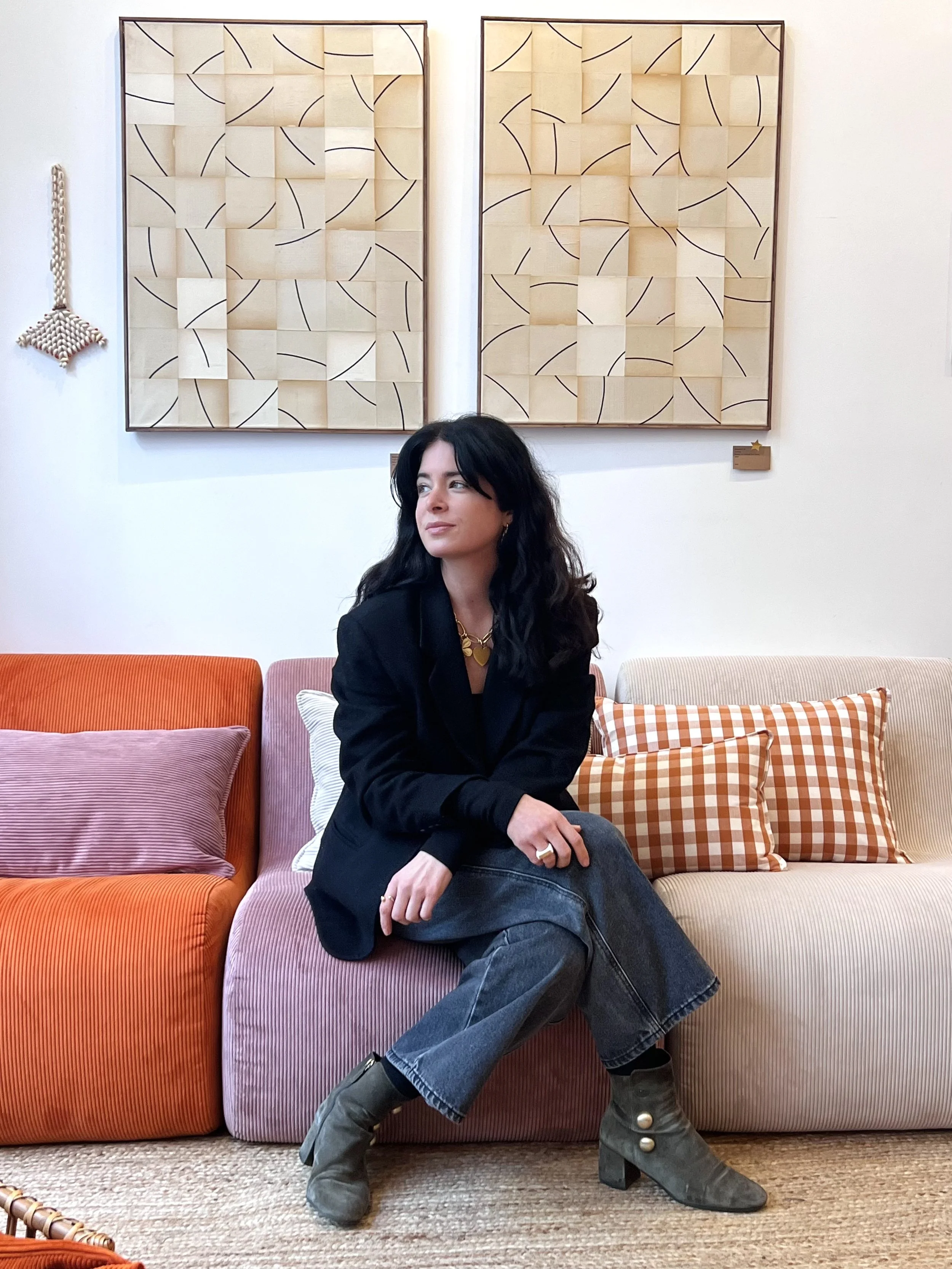 Une femme assise sur un canapé multicolore, avec un fond composé de deux tableaux géométriques en bois suspendus sur un mur blanc.