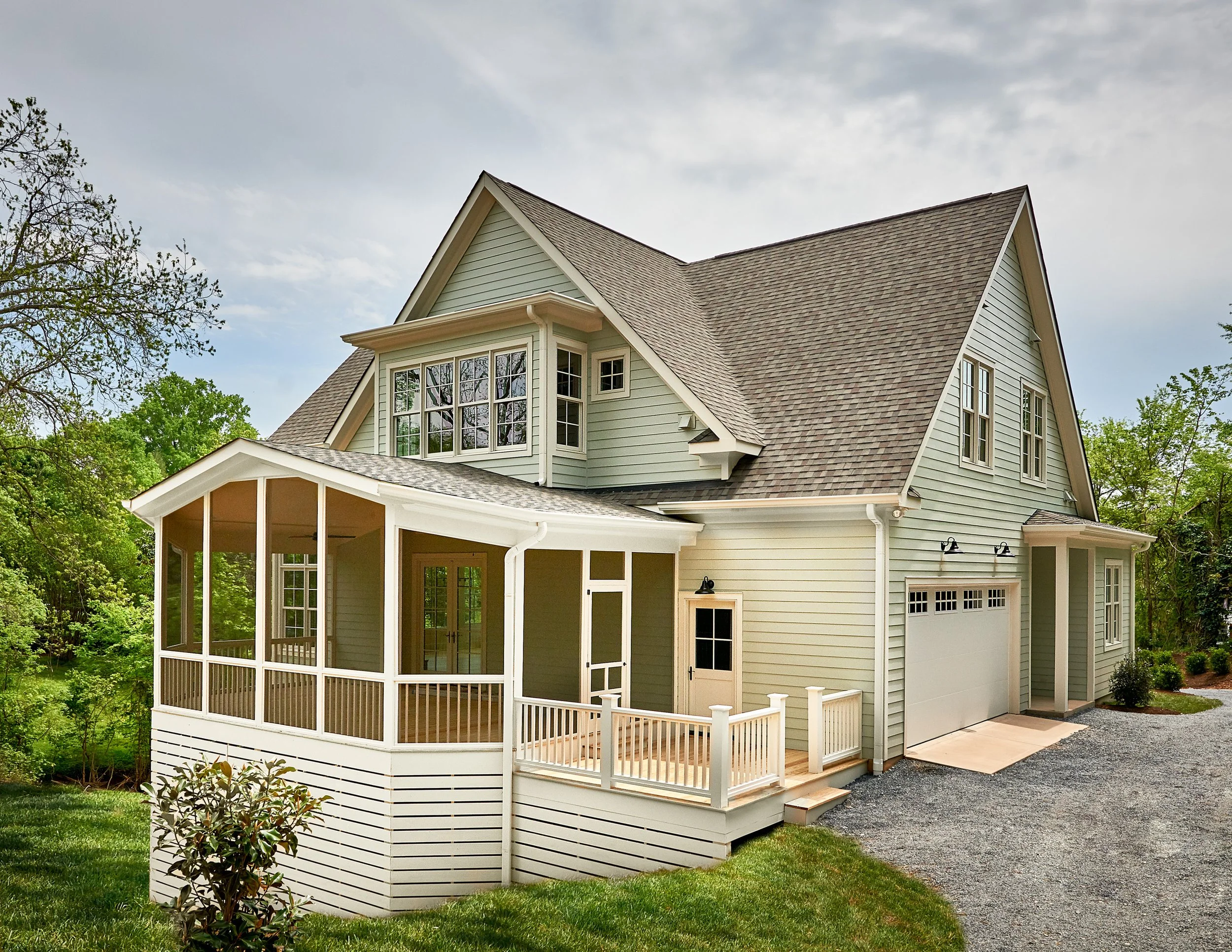 A two-story house with light green siding, a brown shingle roof, and a spacious screened-in porch with white railing and trim, surrounded by green trees and a gravel driveway.