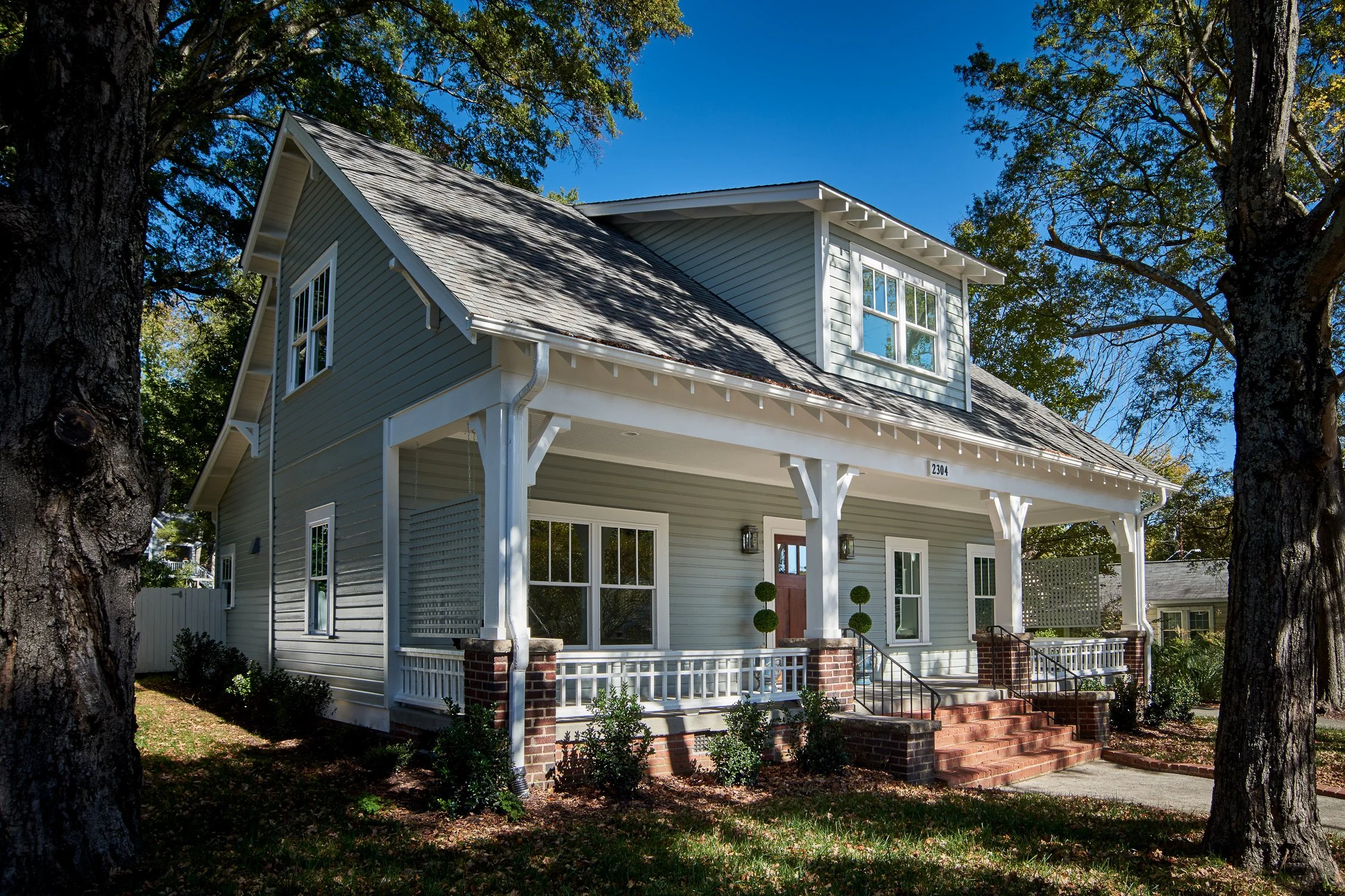 A two-story house with gray siding, white trim, brick steps, and a front porch with decorative posts, situated in a tree-lined neighborhood on a sunny day.