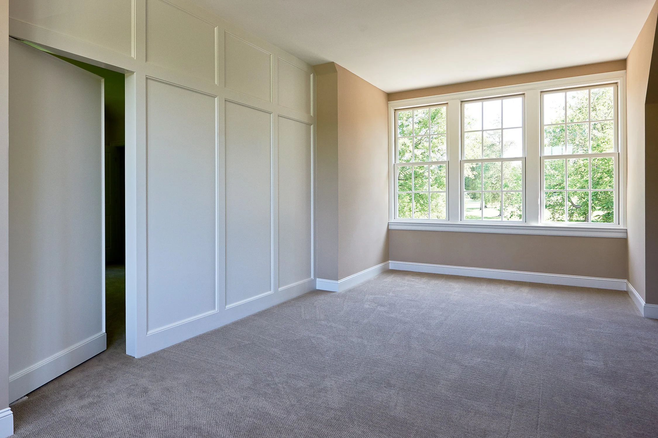Empty beige-walled room with a large window showing green trees outside, white trim, and a beige carpet.