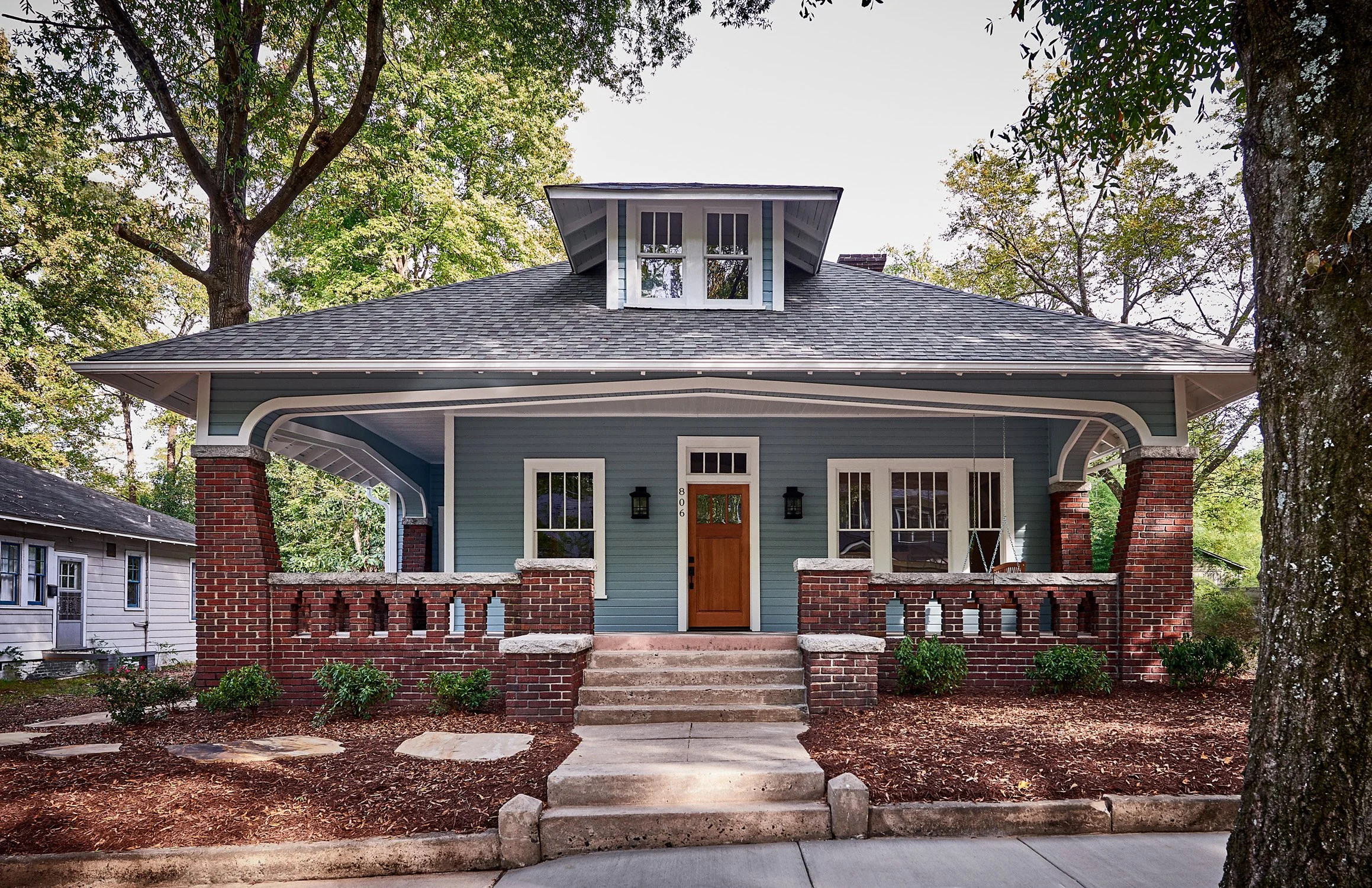 Front view of a charming blue house with a porch, brick railing, and steps leading to the door, surrounded by trees and greenery.