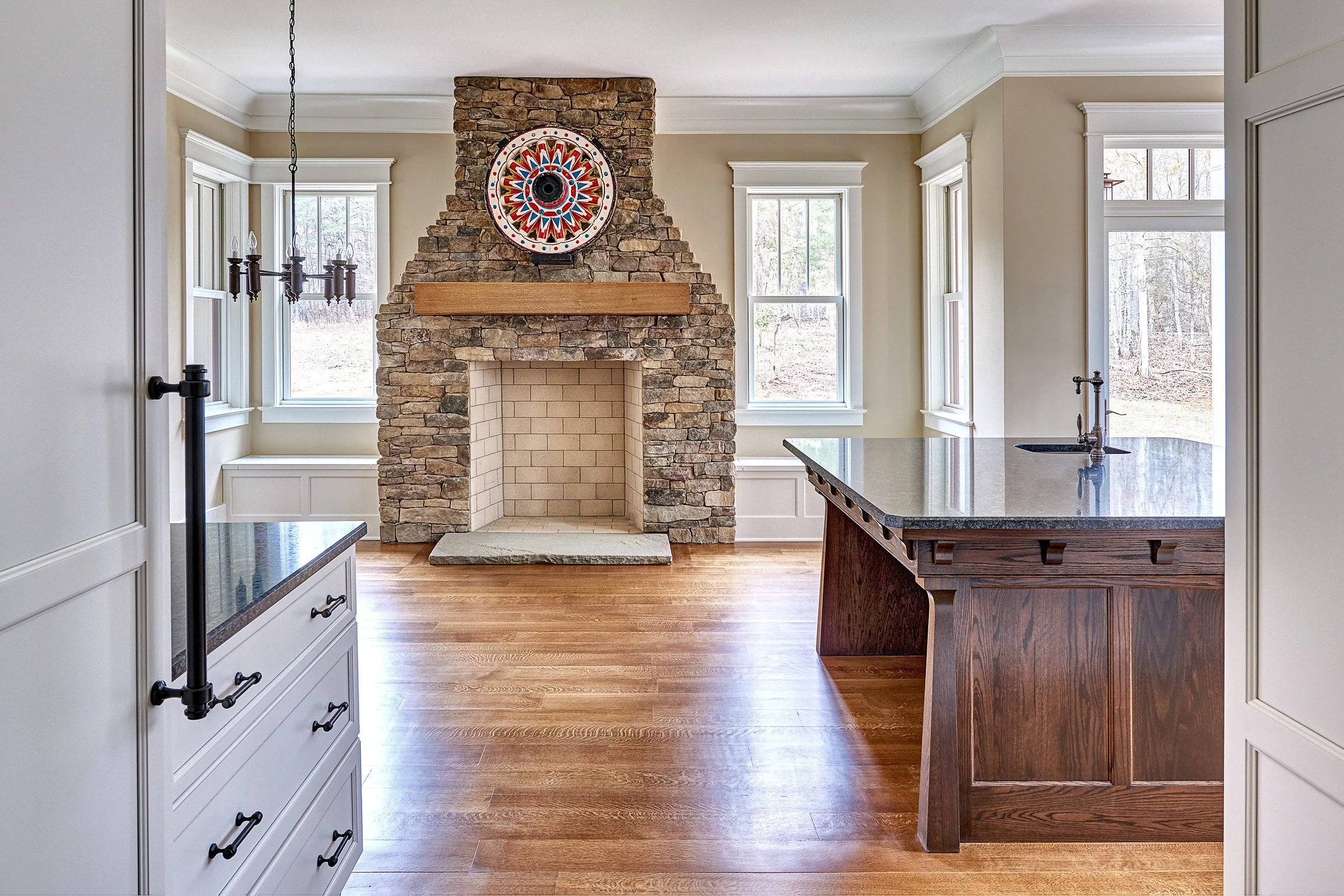 A room with a stone fireplace, wooden mantel, and a colorful decorative plate above the mantel. The room has three windows with white trim, hardwood flooring, and a kitchen island with a dark granite countertop and a sink. There's also a white cabine