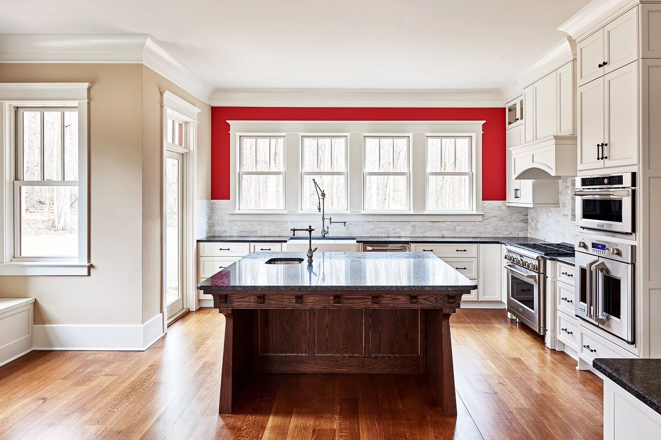 Modern kitchen with white cabinets, a red accent wall, and a central wooden island with a granite countertop. Four large windows above the sink let in natural light, and there are stainless steel appliances including a double oven and a stove.