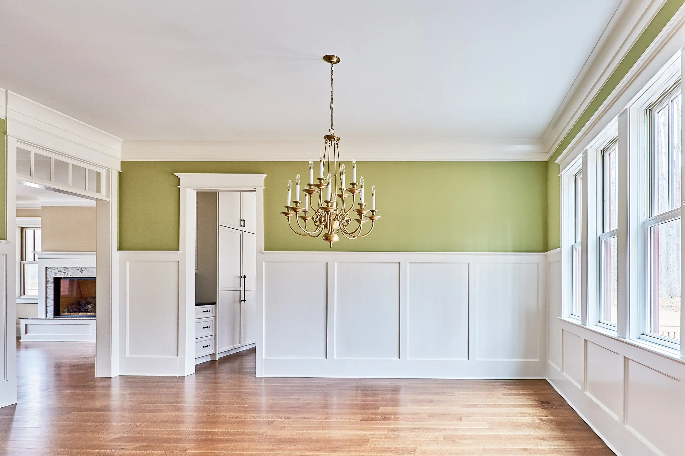 Empty dining room with green wall, white wainscoting, and large windows, with a chandelier hanging from the ceiling.
