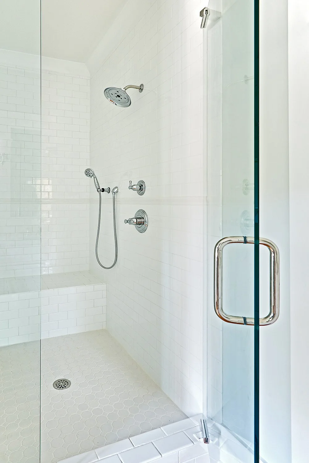 Clean, modern shower with white tiles, a rainfall showerhead, a handheld shower, control knobs, and a glass door with a metal handle.