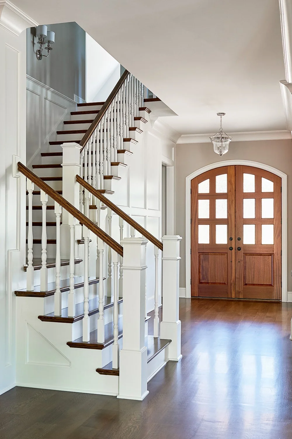 Interior view of a home with a wooden front door, staircase with white balusters and wooden handrails, hardwood floors, wall paneling, and a hanging light fixture.