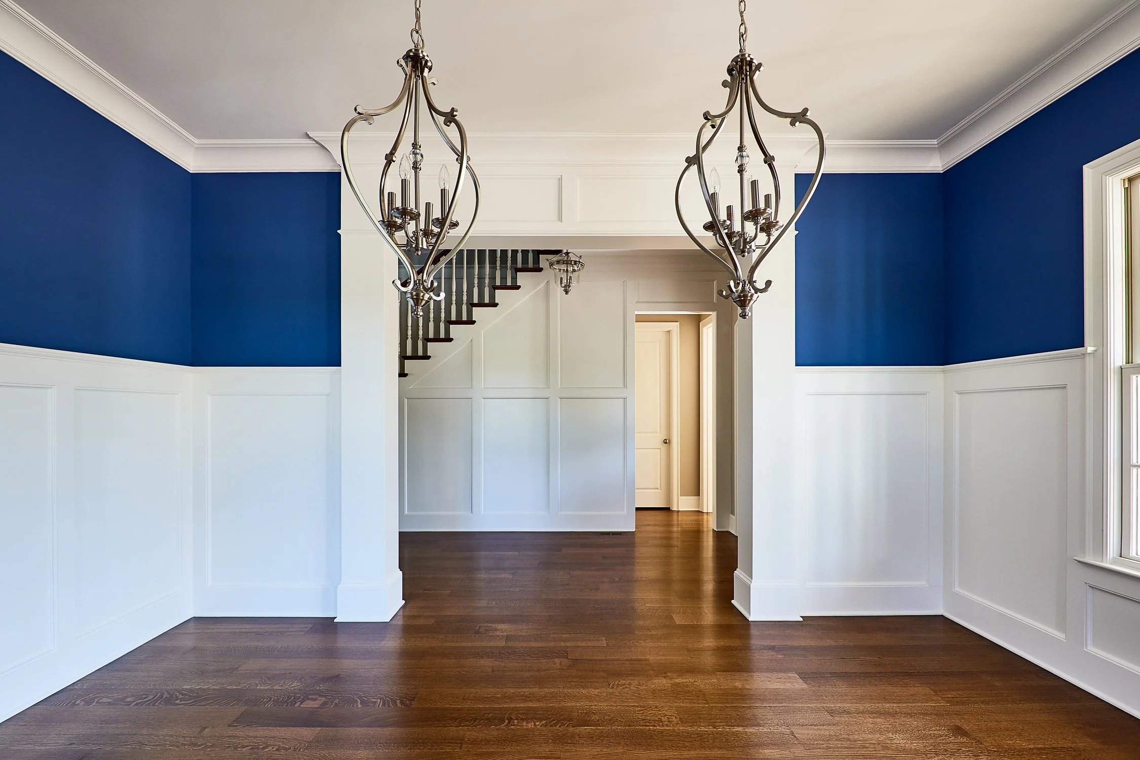 Empty dining room with blue and white walls, brown hardwood floors, two chandeliers, staircase, and door.