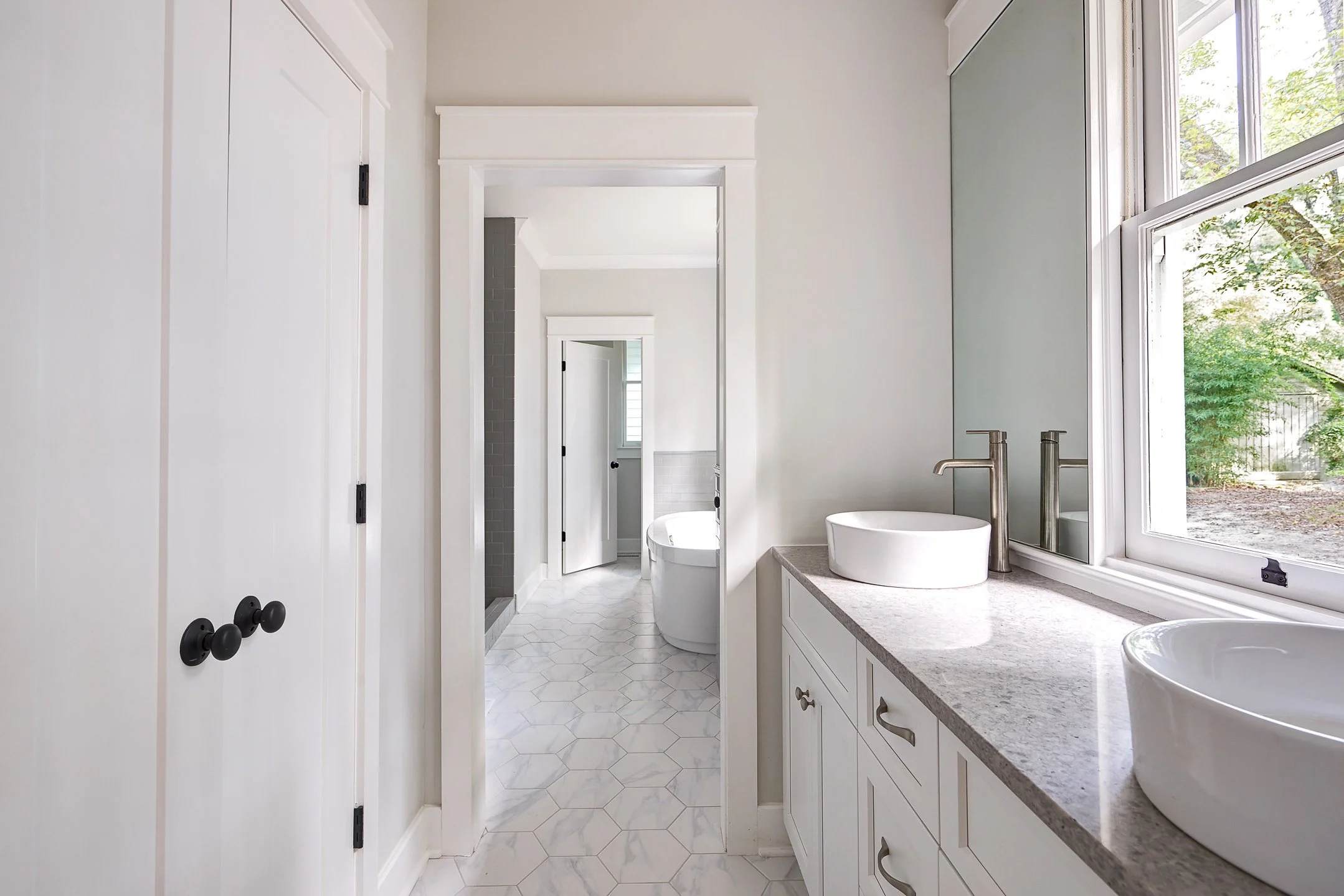 Bright and modern bathroom with white vanity, two vessel sinks, a large window showing greenery outside, and a view into a room with a bathtub and hexagonal floor tiles.
