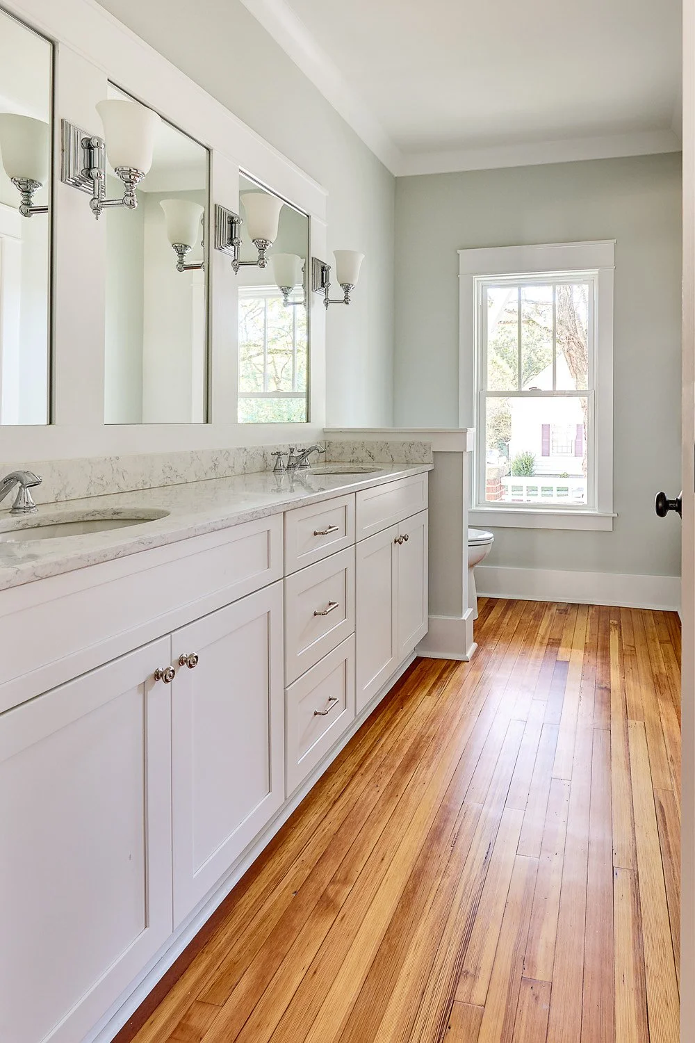 A bright bathroom with a white double vanity with a marble countertop, three mirrors, and four light fixtures above each mirror. There is a window letting in natural light, showing trees and a house outside, with a wooden floor and light-colored wall